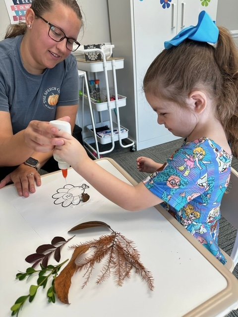 A young girl with a blue bow in her hair and a colorful cartoon shirt is gluing dried leaves and flowers onto a drawing of a rabbit on a table, with a woman helping her work on occupational therapy goals.