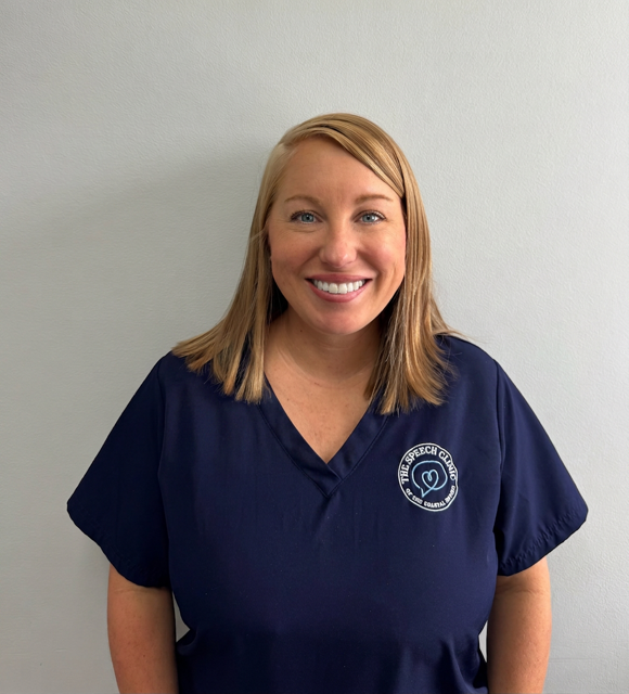 A woman with shoulder length blonde hair smiling, wearing navy scrubs with a logo on the chest against a plain white wall. Billing Specialist at The Speech Clinic.