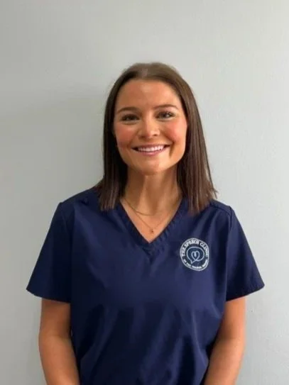 A woman in a navy blue medical scrub top standing in front of a plain light-colored wall, smiling at the camera. Therapist at The Speech Clinic of the Coastal Empire.