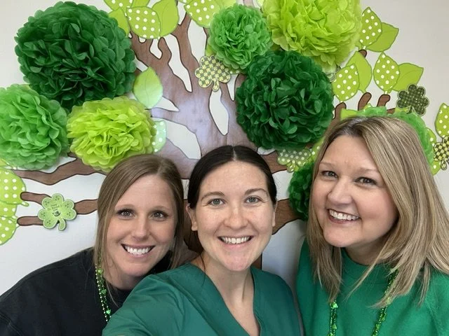 Three women smiling in front of a paper tree with green paper pom-pom leaves and decorated with shamrocks, possibly for St. Patrick's Day celebration. They work together at The Speech Clinic of the Coastal Empire.