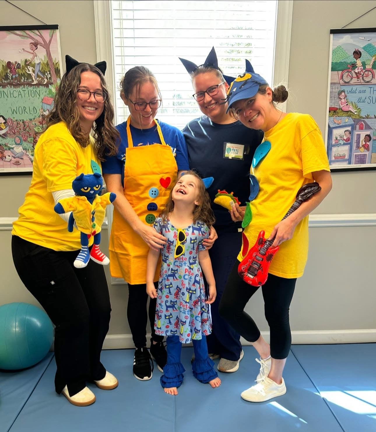 Group of five smiling women and a young girl in a playful, colorful room with posters on the wall, dressed in costumes with cat ears and accessories.  Therapists with The Speech Clinic of the Coastal Empire in Savannah GA.