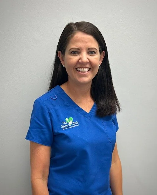 A woman with long dark hair wearing blue medical scrubs, standing against a plain light gray wall, smiling at the camera. A therapist with The Speech Clinic of the Coastal Empire in Savannah GA.