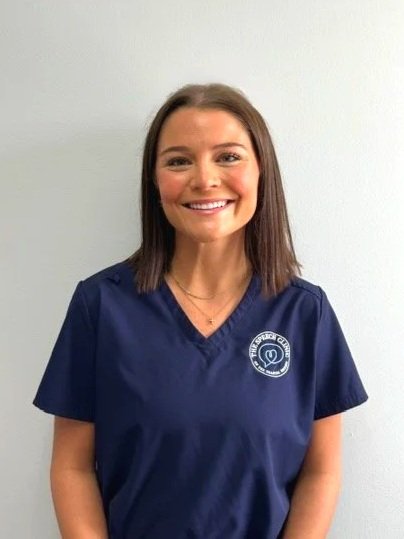 A smiling woman with shoulder-length brown hair wearing navy medical scrubs with an emblem on the chest, standing against a plain white wall. Therapist at The Speech Clinic of the Coastal Empire.
