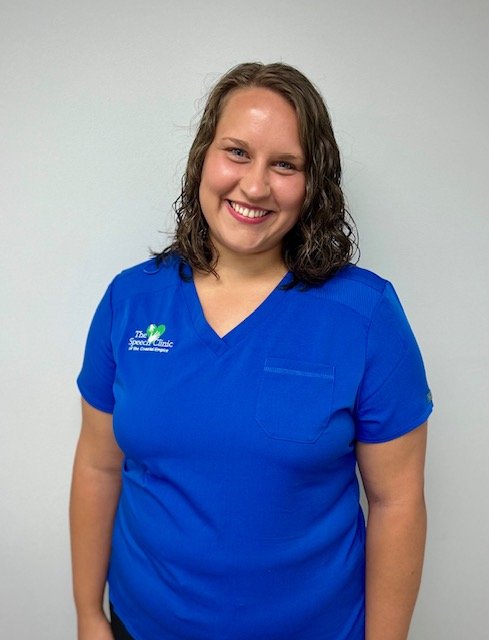 A young woman with curly brown hair wearing a blue medical scrub top with a logo on the chest, smiling at the camera.  A therapist with The Speech Clinic of the Coastal Empire in Savannah GA.