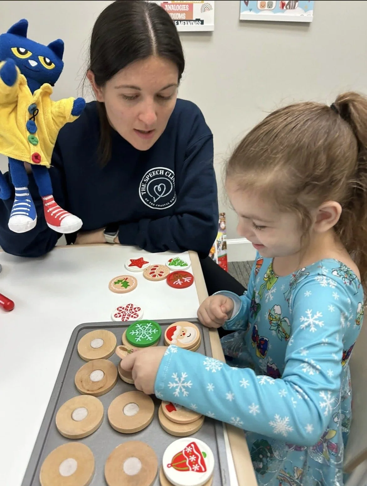A woman and a girl sitting at a table playing with holiday-themed wooden cookie toys. They are engaging with decorated cookie-shaped pieces, while working on speech therapy goals.