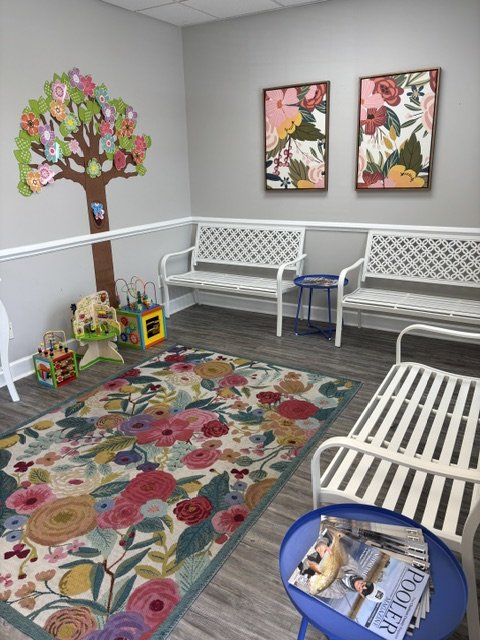 Colorful children's waiting room with floral rug, white benches, toy shelves, wall art, and a tree mural. A fun entrance into The Speech Clinic of the Coastal Empire.