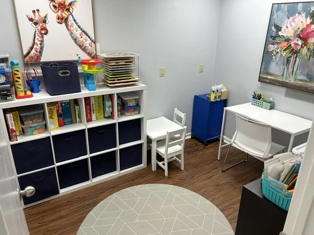 A small playroom with shelves filled with books and toys, a white table and chair, a blue cabinet, and wall art featuring giraffes and flowers. A therapy room at The Speech Clinic of the Coastal Empire.