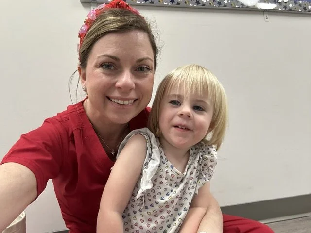 A woman and a young girl sitting together, smiling for a selfie, in a room with a white wall and colorful star decorations in the background. Working on speech therapy goals.