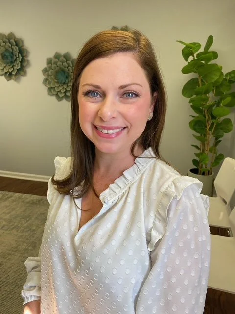 A woman with brown hair and blue eyes smiling, wearing a white blouse with ruffled shoulders, standing in a room with green plants and decorative wall art. Speech Pathologist and owner of The Speech Clinic of the Coastal Empire.