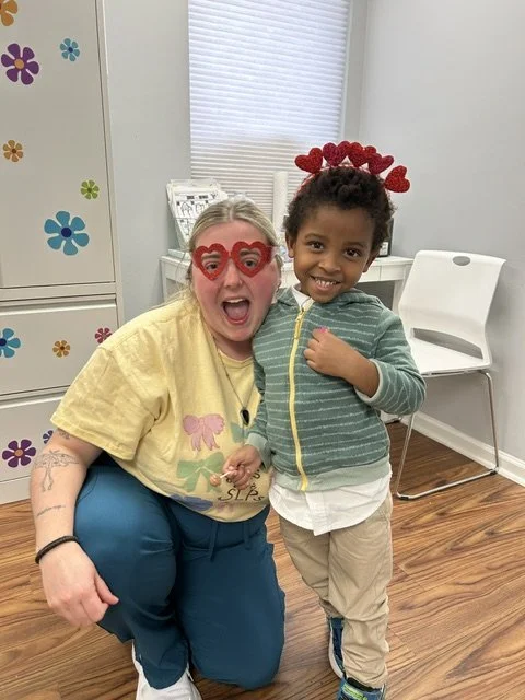 A woman and a young boy celebrating Valentine's Day indoors. The woman is wearing red heart-shaped glasses and smiling, while the boy is wearing a red heart headband and smiling. They just finished their speech therapy session.