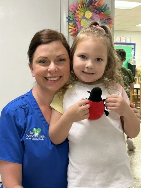 A woman in a blue shirt smiling with a young girl holding a plush ladybug toy in a classroom. Working on speech therapy goals.