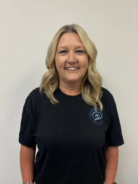 A woman with blonde hair wearing a black shirt with a logo, standing against a plain white wall and smiling. Intake Specialist at TSCCE.