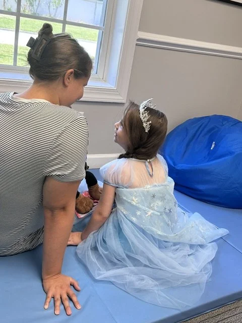 A woman and young girl sitting on a floor mat, facing each other, with a window behind them showing a grassy yard. The girl is dressed like a princess with a blue ball gown and tiara. They are in an Occupational Therapy session.
