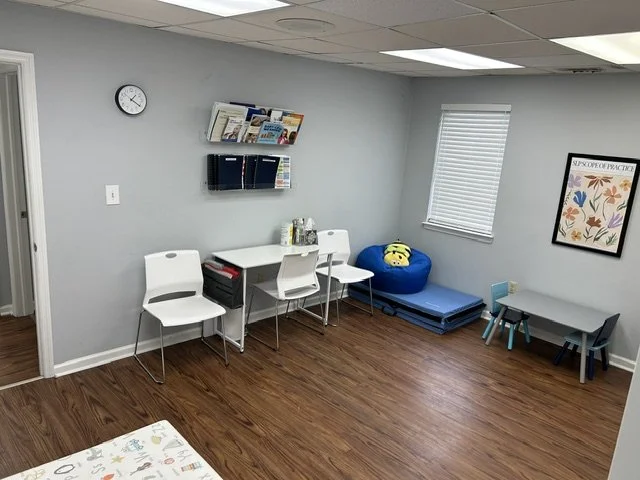 Empty therapy or counseling room with chairs, a small table, a framed artwork, and plush toys on a blue bench. A therapy room at The Speech Clinic of the Coastal Empire.