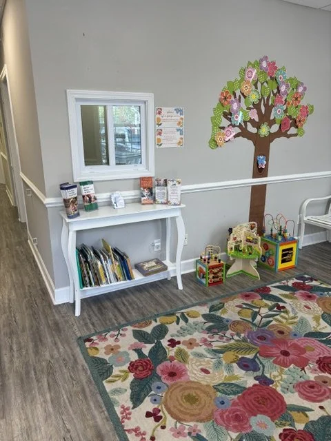 Indoor children’s play area with a colorful floral rug, a white wall with a tree and flower decoration, a small white table with books and pamphlets, and toys including a bead maze. A therapy room at The Speech Clinic of the Coastal Empire.
