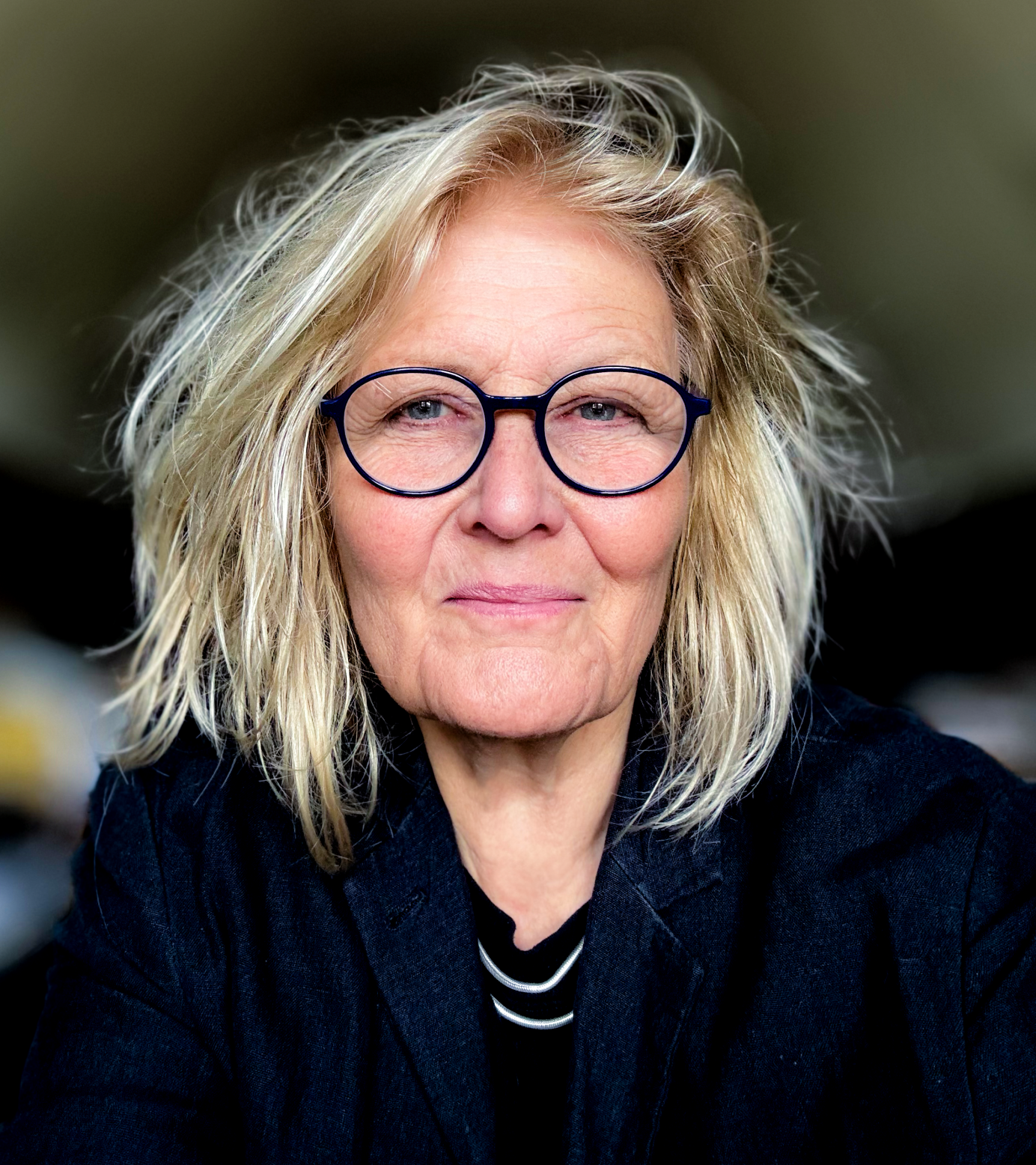 Close-up of older woman with wavy blonde hair, wearing round glasses and a dark jacket, looking directly at the camera.