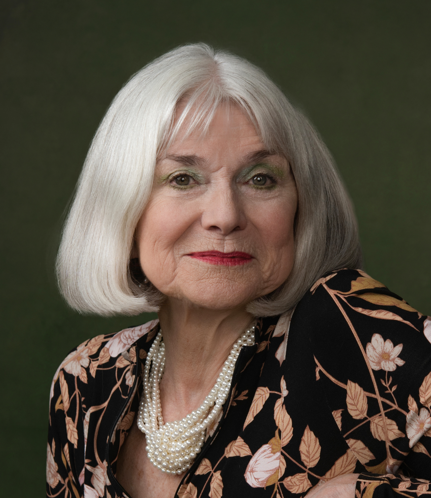 An older woman with shoulder-length silver hair, green eyeshadow, red lipstick, wearing a black jacket with a floral pattern and a pearl necklace, against a dark green background.