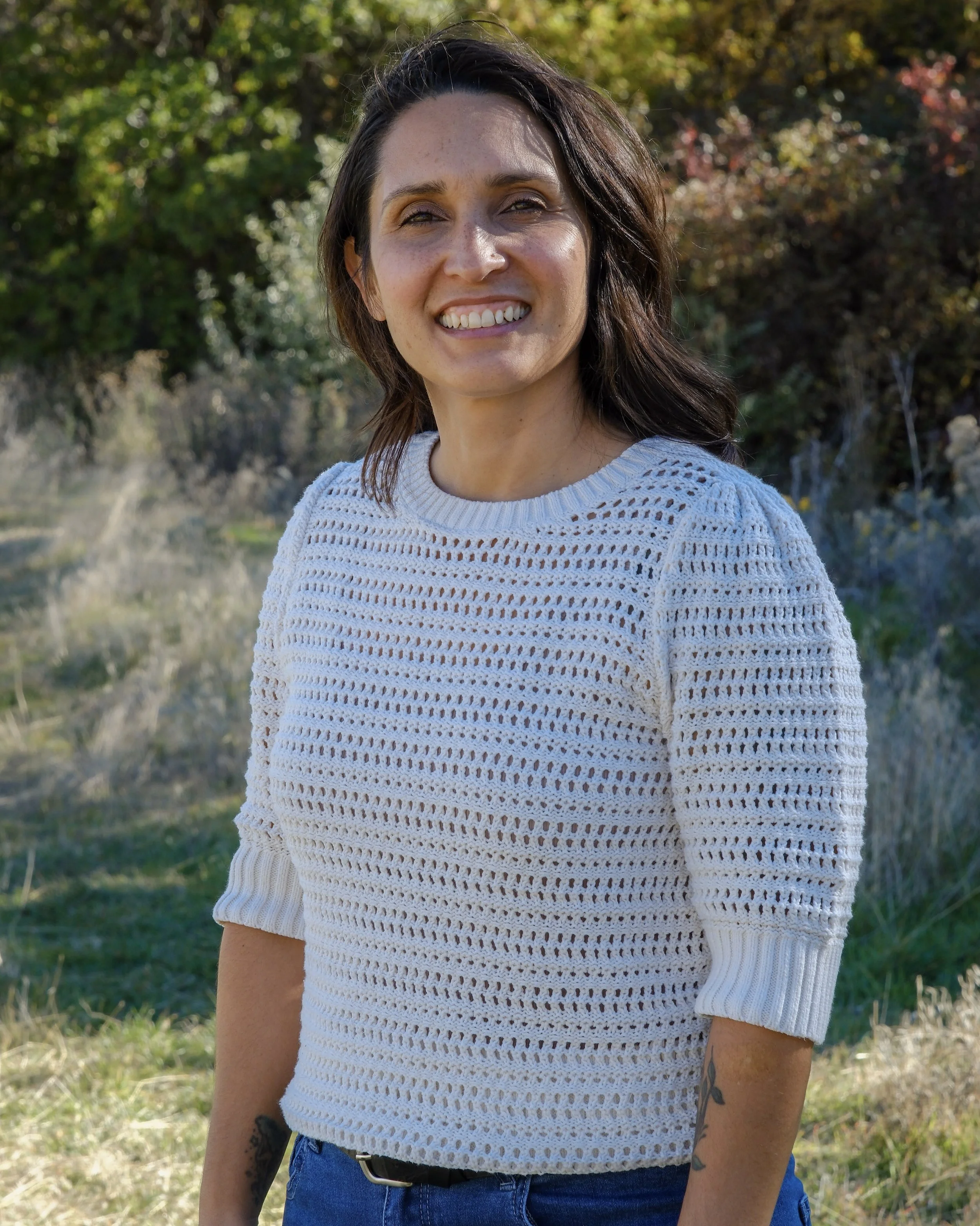 A woman with shoulder-length brown hair smiling outdoors, wearing a white open-knit sweater and blue jeans, with trees and grass in the background.