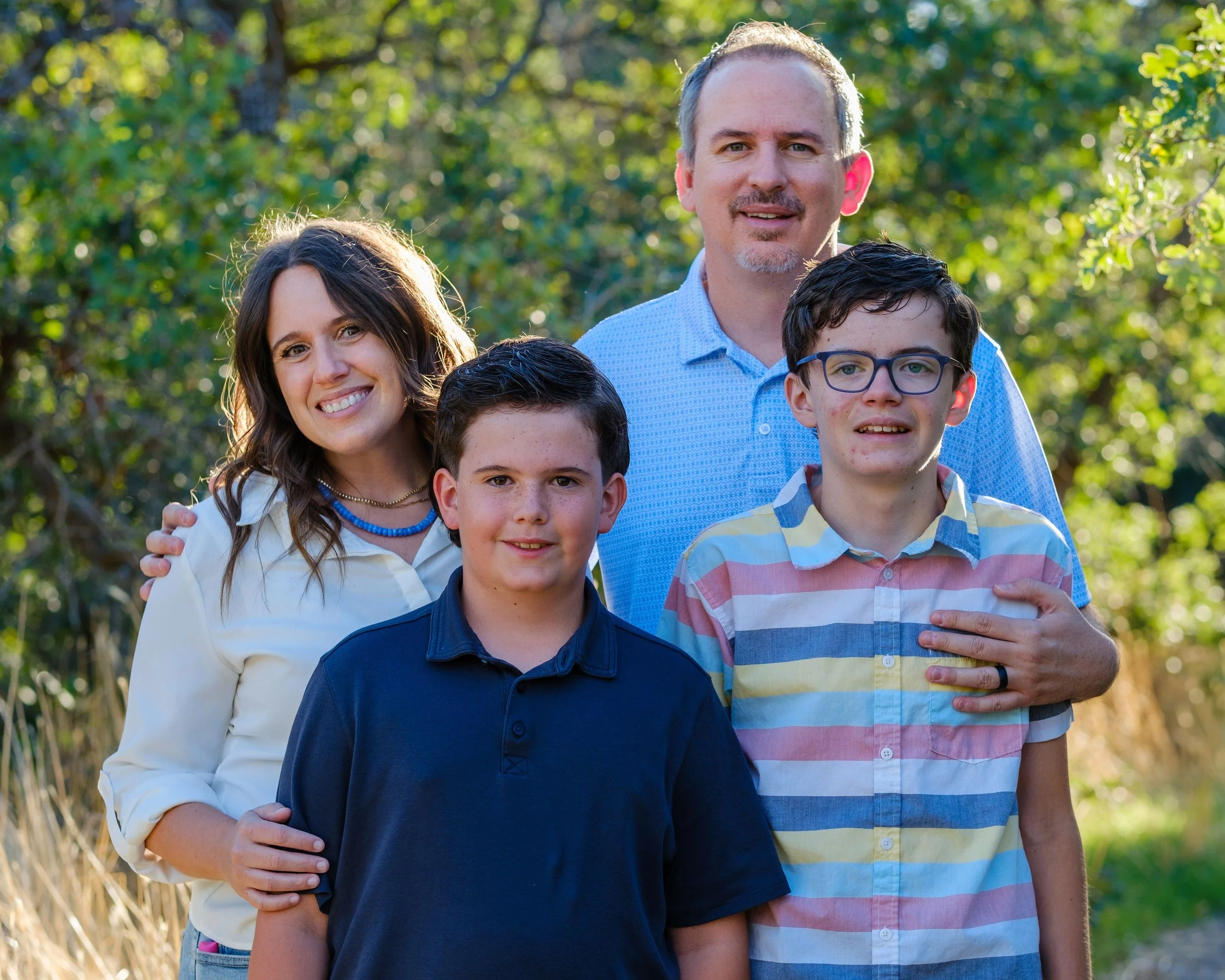 A family of five standing outdoors in a sunny park with green trees and grass in the background. The family includes a woman, a man, and three children. They are all smiling and appear happy.