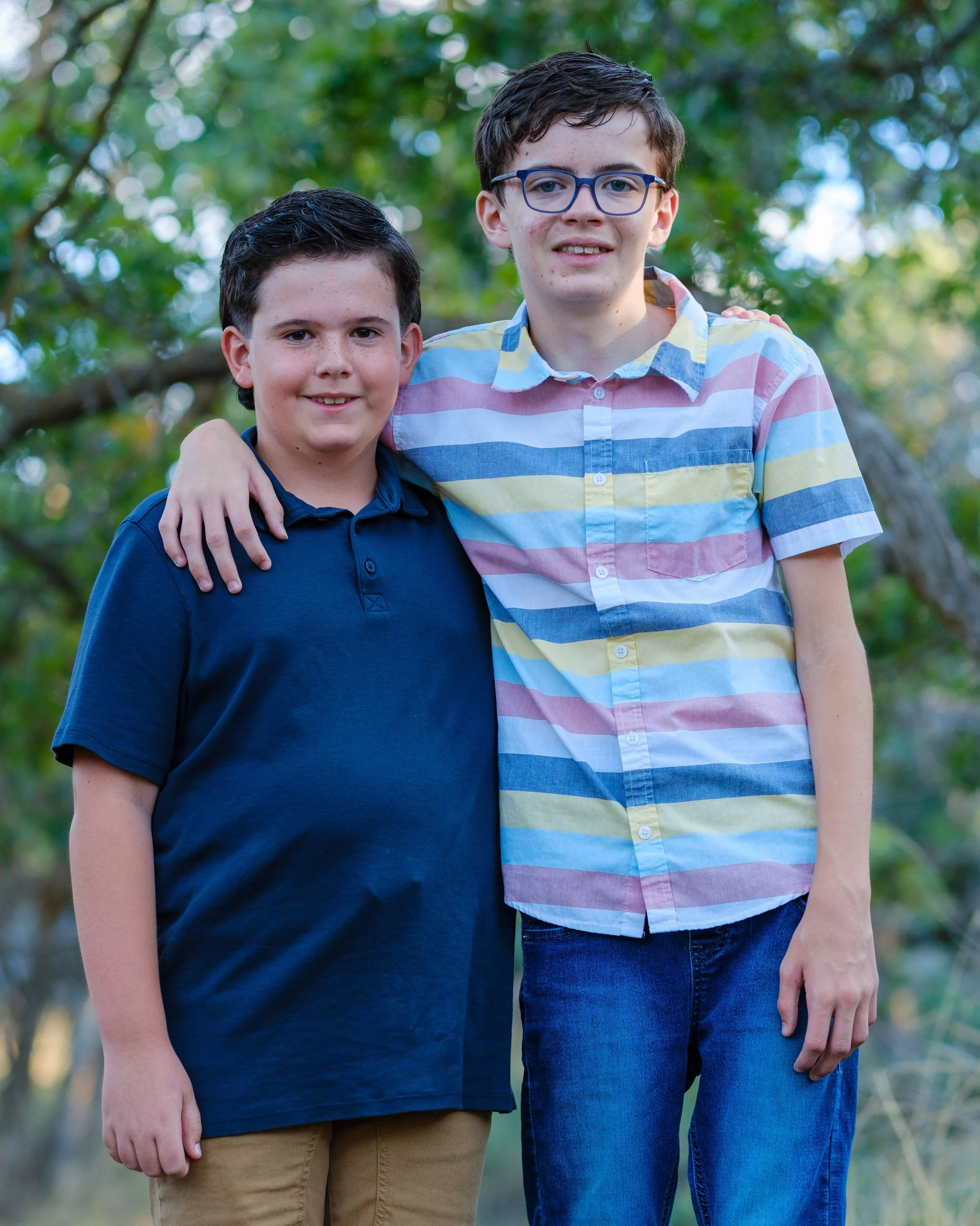 Two boys standing outdoors with trees in the background, one with glasses and both smiling, one wearing a striped shirt and the other a dark polo shirt.