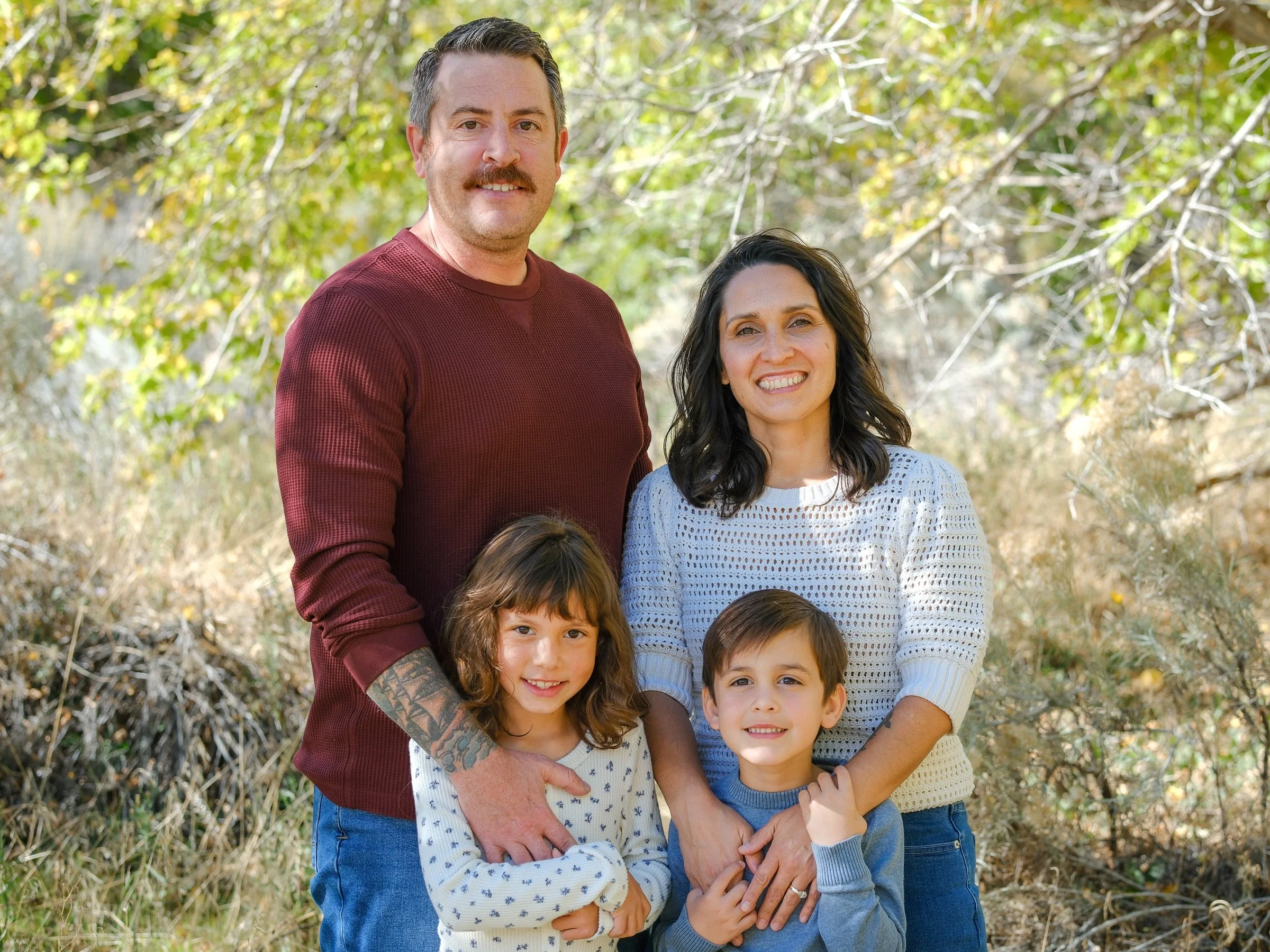 A family of five including two women, one man, and two children standing outdoors in a natural setting with trees and green foliage.