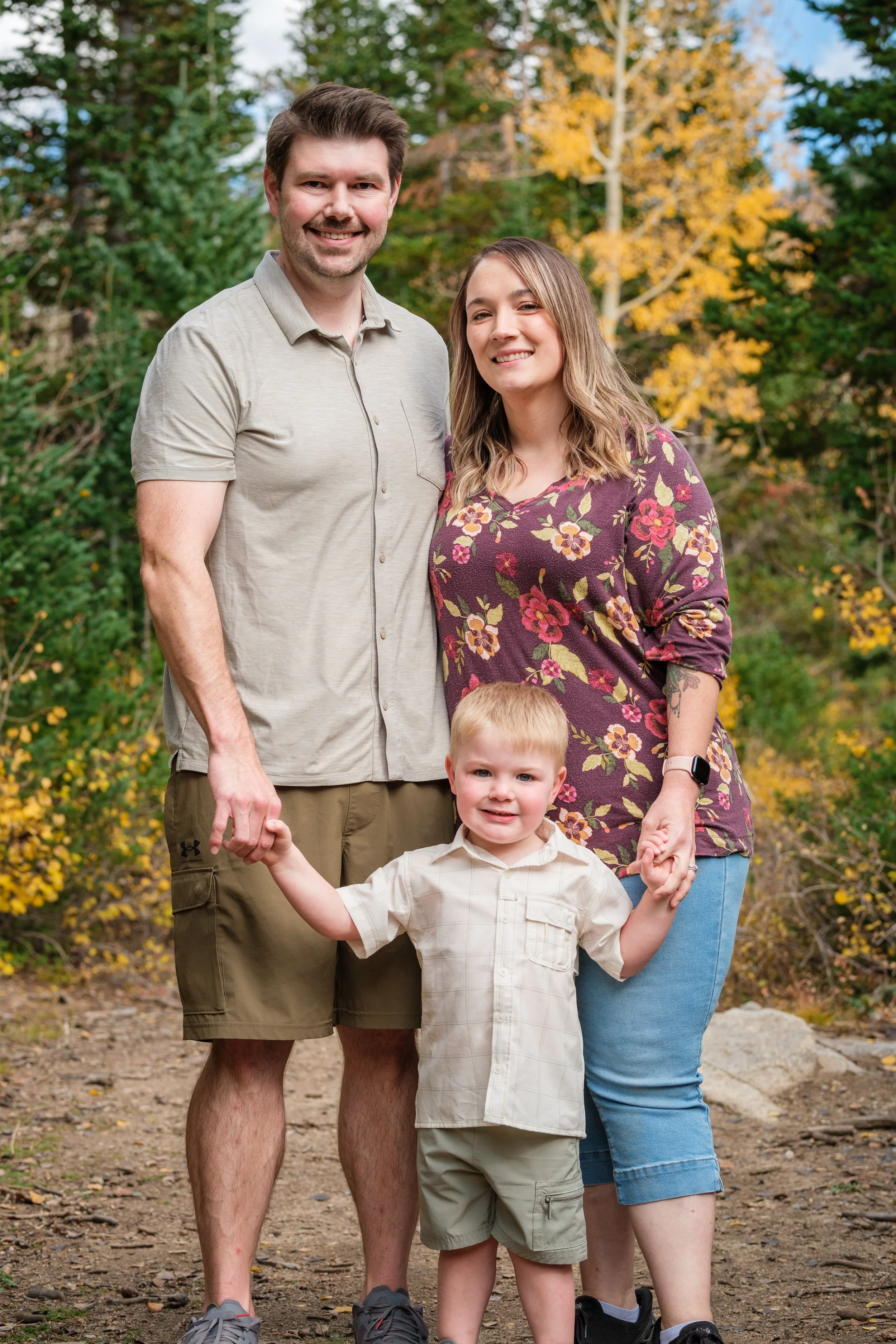 A family of four standing outdoors with trees and fall foliage in the background. The father is on the left, smiling, holding hands with the young boy in the foreground. The mother is on the right, also smiling, holding hands with the young boy and s