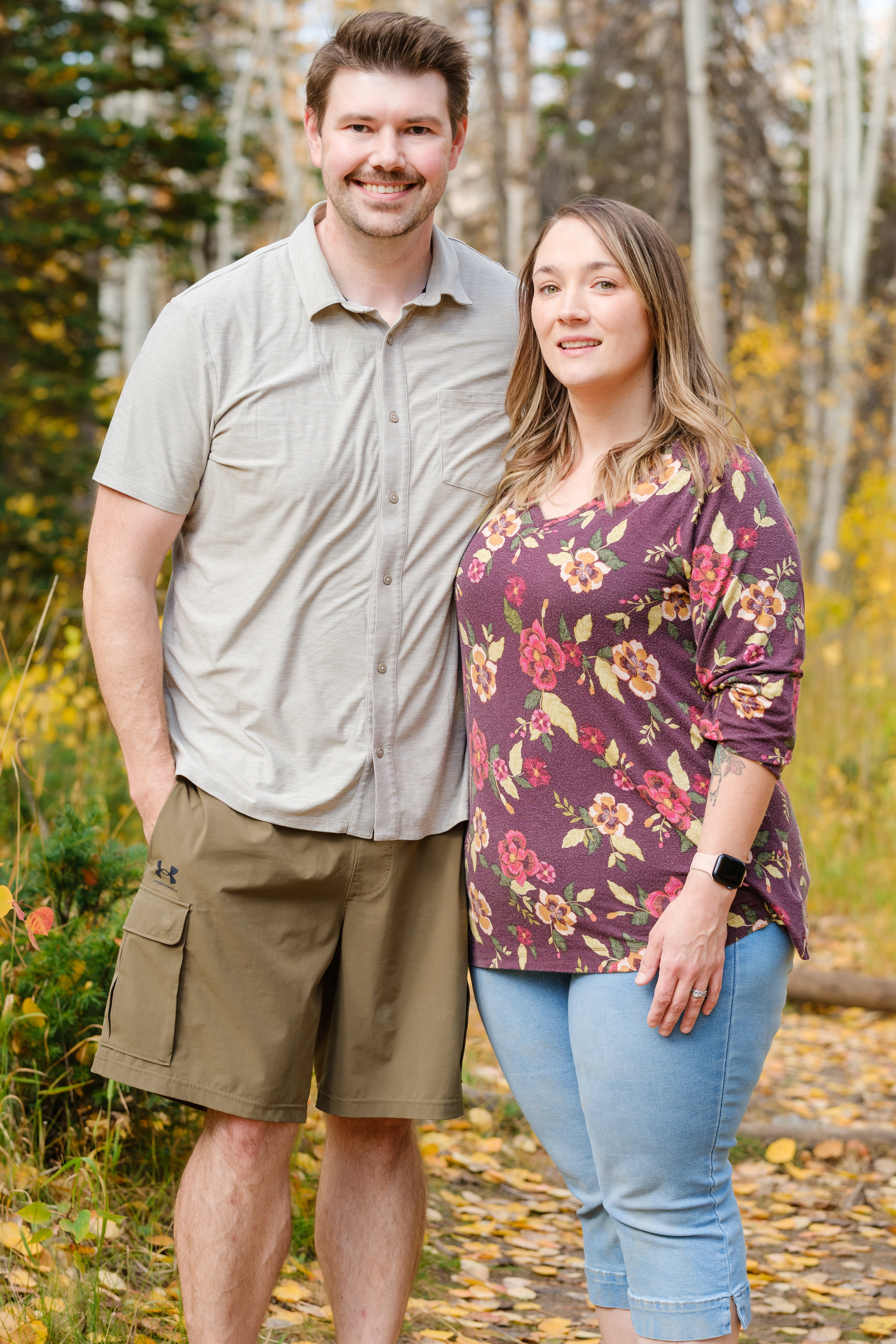 A smiling man and woman standing outdoors on a fall day among trees with yellow leaves. The man is wearing a light gray short-sleeve button-up shirt and khaki shorts. The woman is dressed in a purple floral long-sleeve shirt and light blue jeans. The