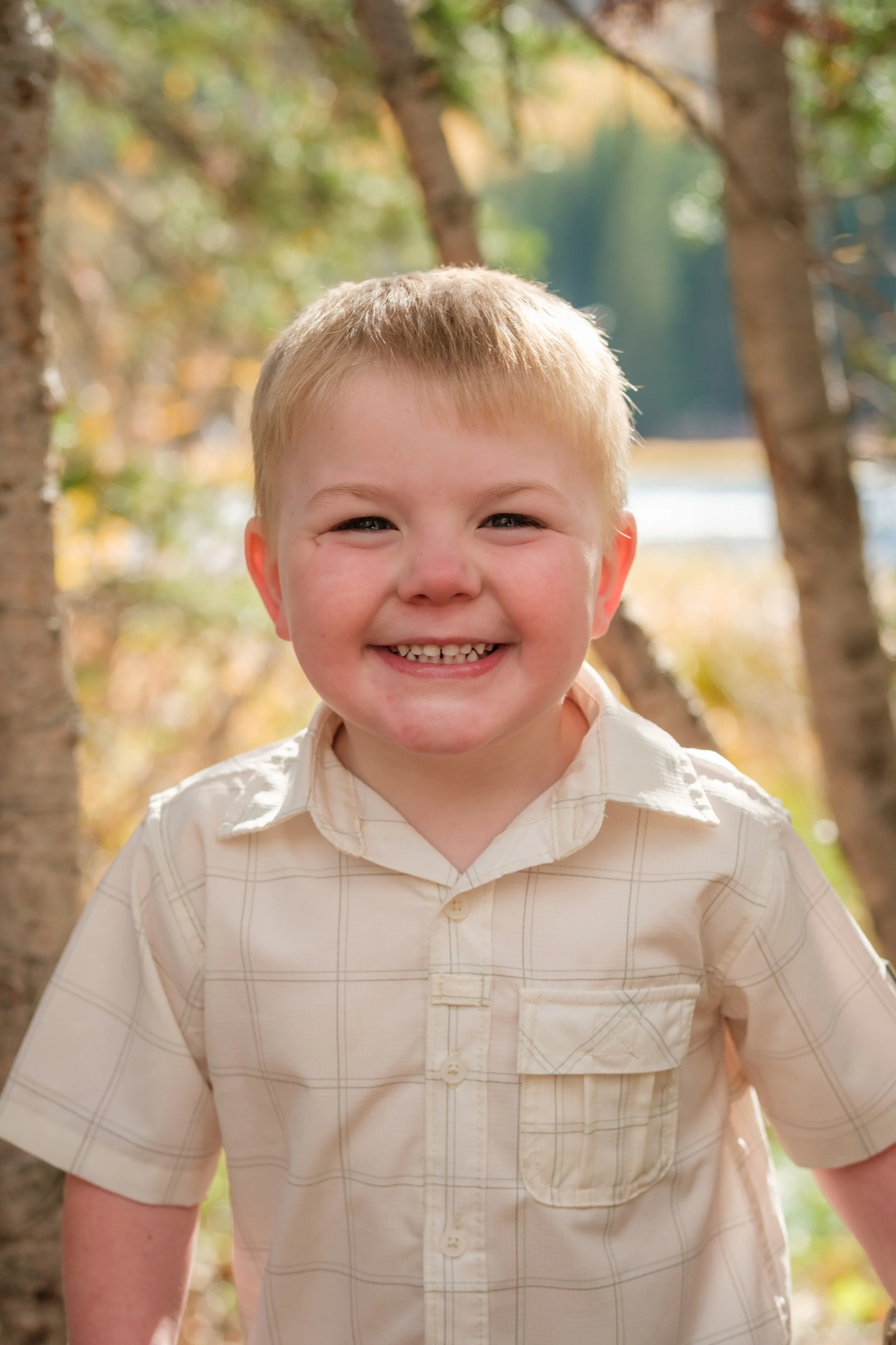 Close-up of a young boy with blond hair and blue eyes smiling outdoors in front of trees with autumn foliage.