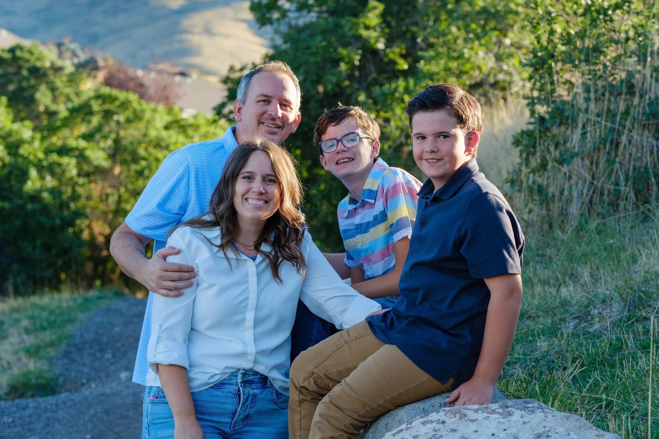 Family of five posing outdoors on a sunny day with trees and hills in the background.