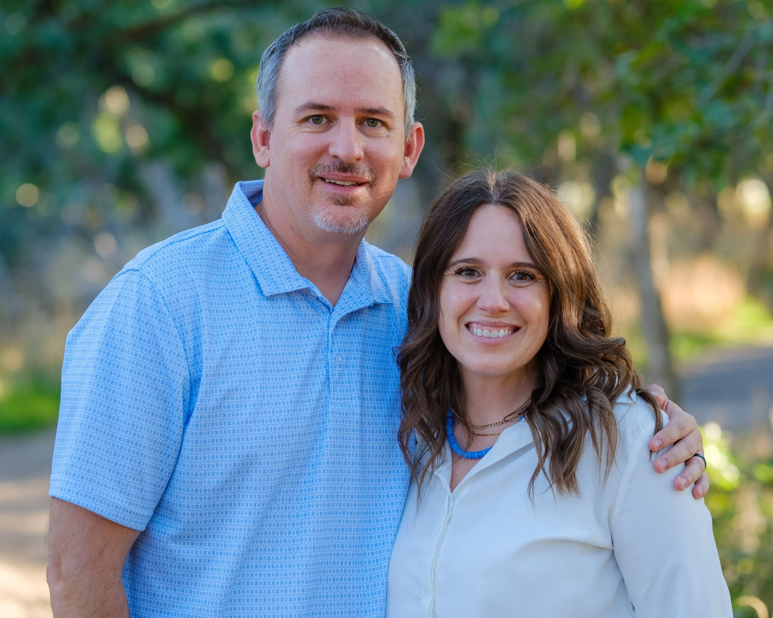 A man with short dark hair and a goatee standing outdoors, smiling, with a woman with long brown hair and a white shirt, smiling, standing next to him, both with greenery in the background during daytime.