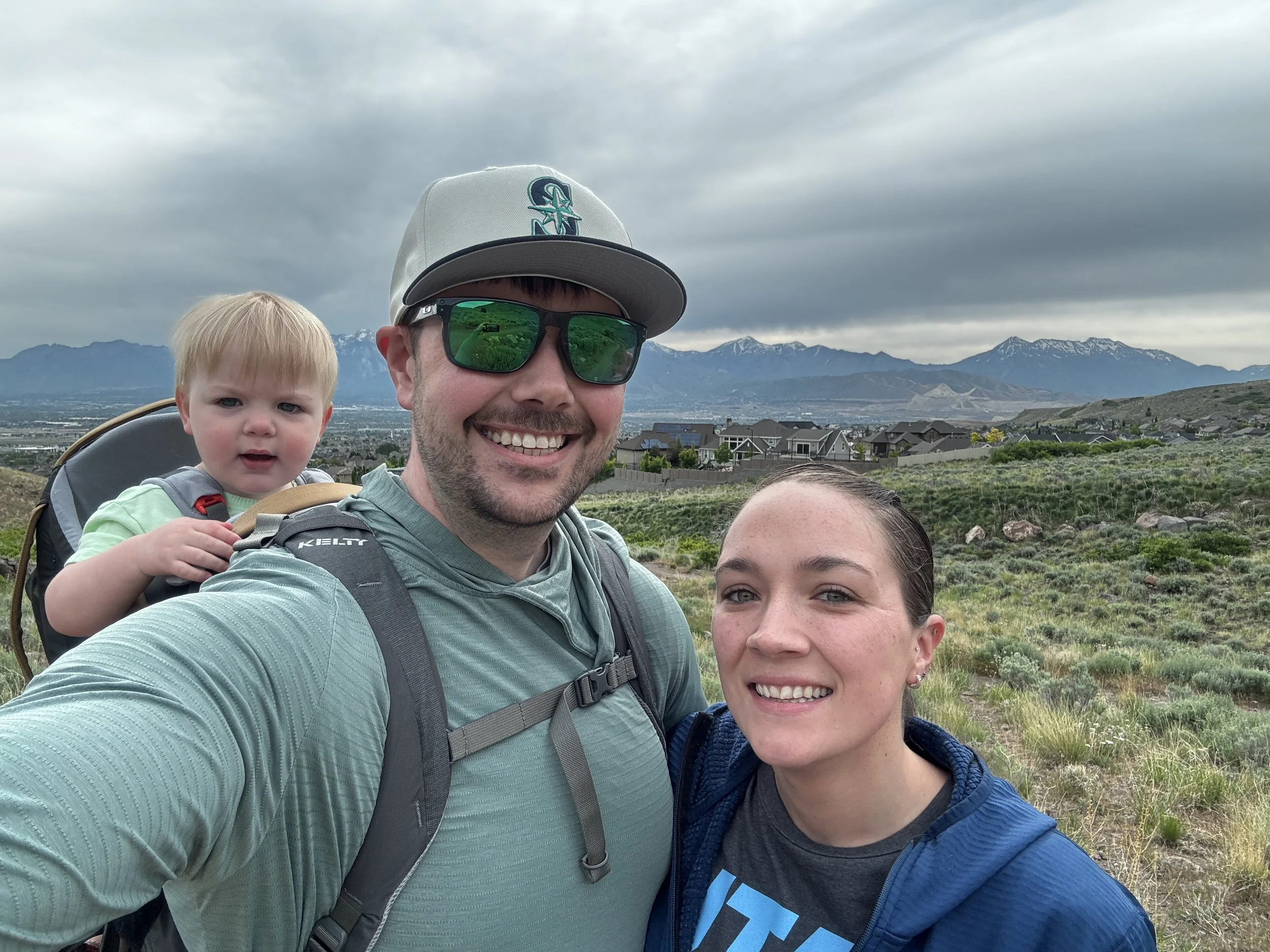 A man with a backpack and sunglasses smiling, a woman with a hoodie, and a young child with blonde hair in a backpack carrier, taking a selfie during a hike with a backdrop of mountains, a residential area, and a cloudy sky.