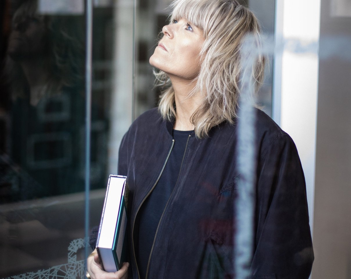 Femme aux cheveux blonds, portant une veste noire, regardant vers le haut et tenant un livre.