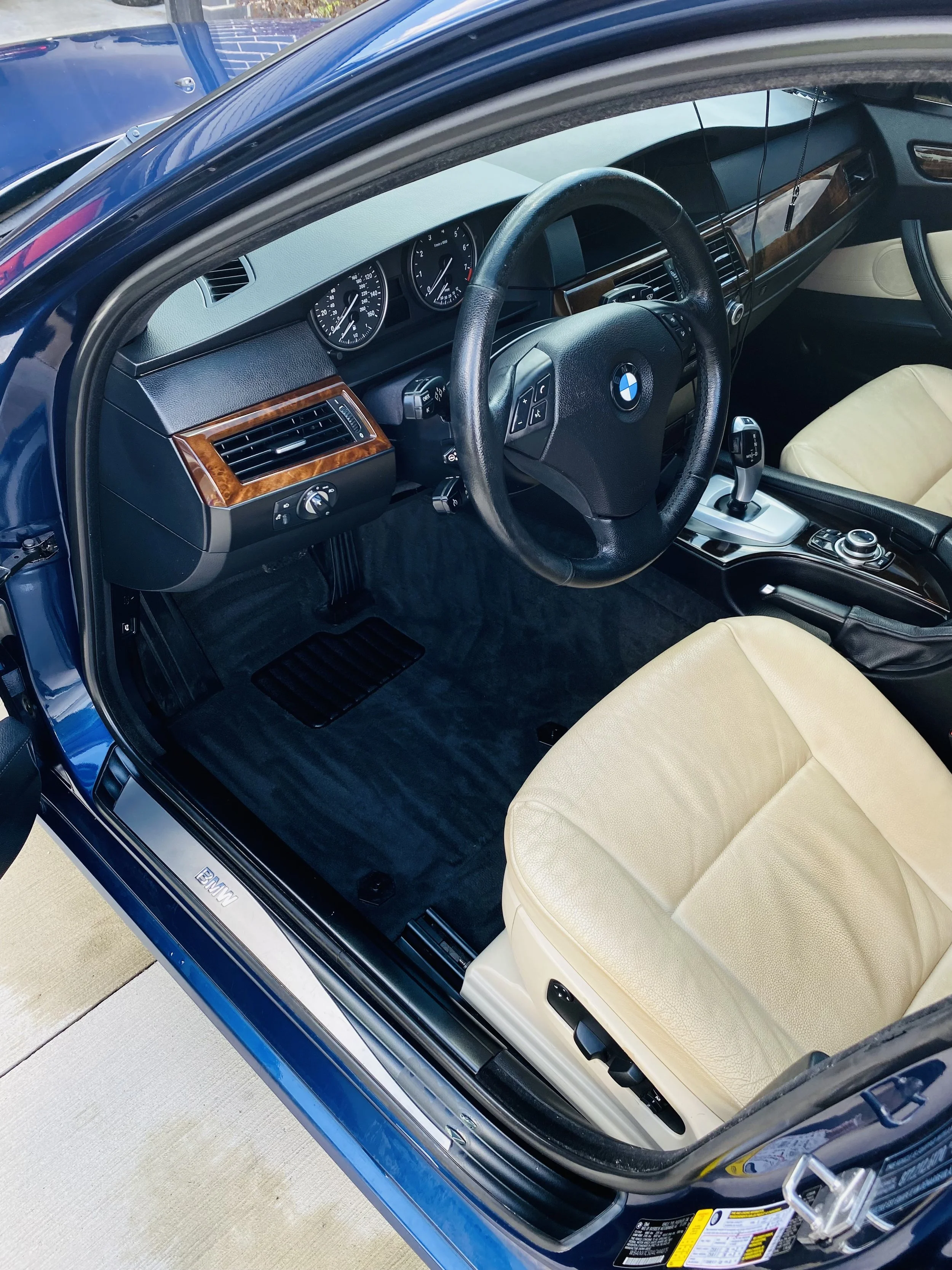 Interior of a blue BMW vehicle showing the dashboard, steering wheel, beige leather seats, gear shift, and part of the driver's side door.