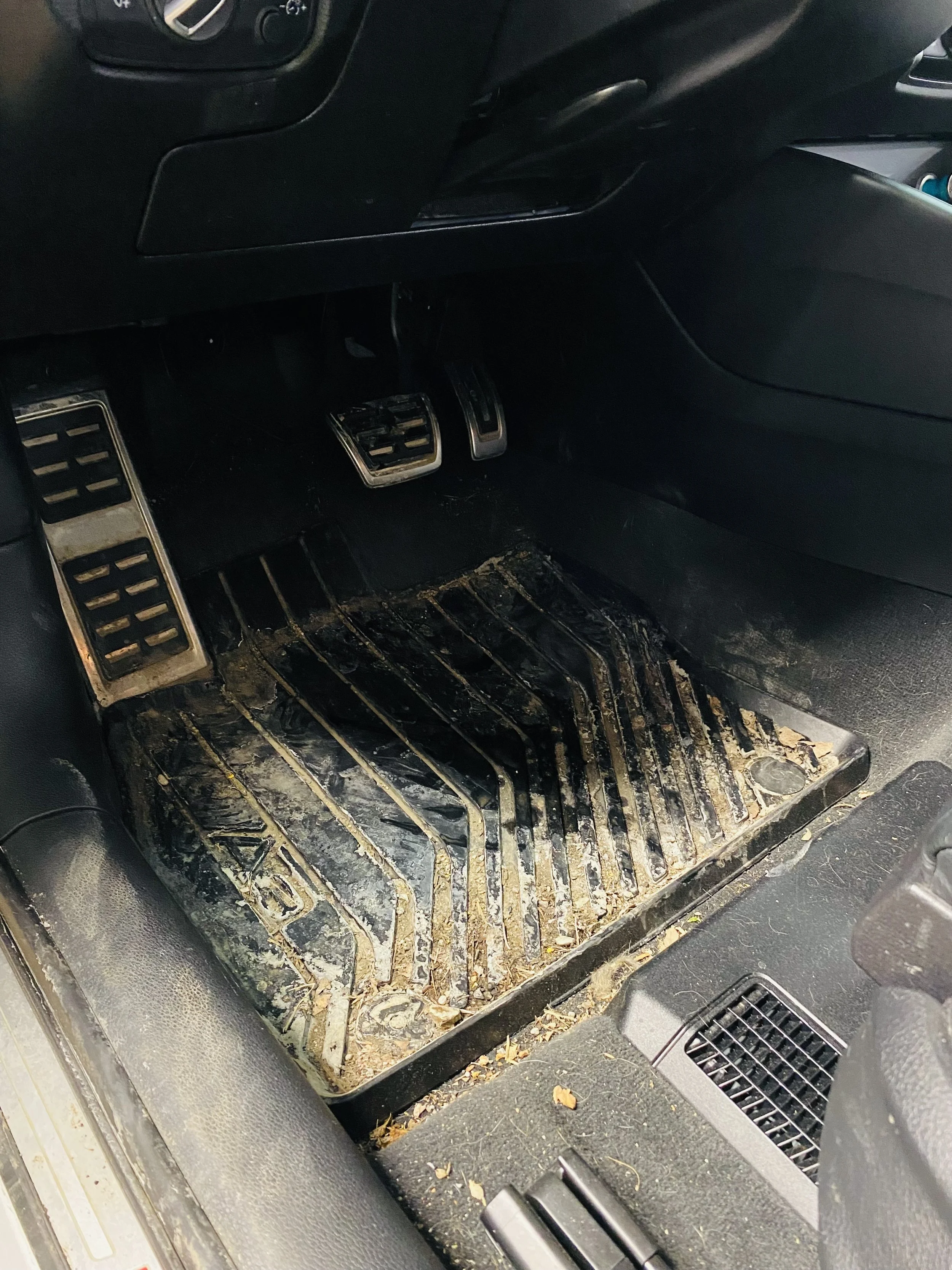 Dirty car floor mat with dirt, leaves, and debris, near the driver's side footwell, with the accelerator and brake pedals visible.