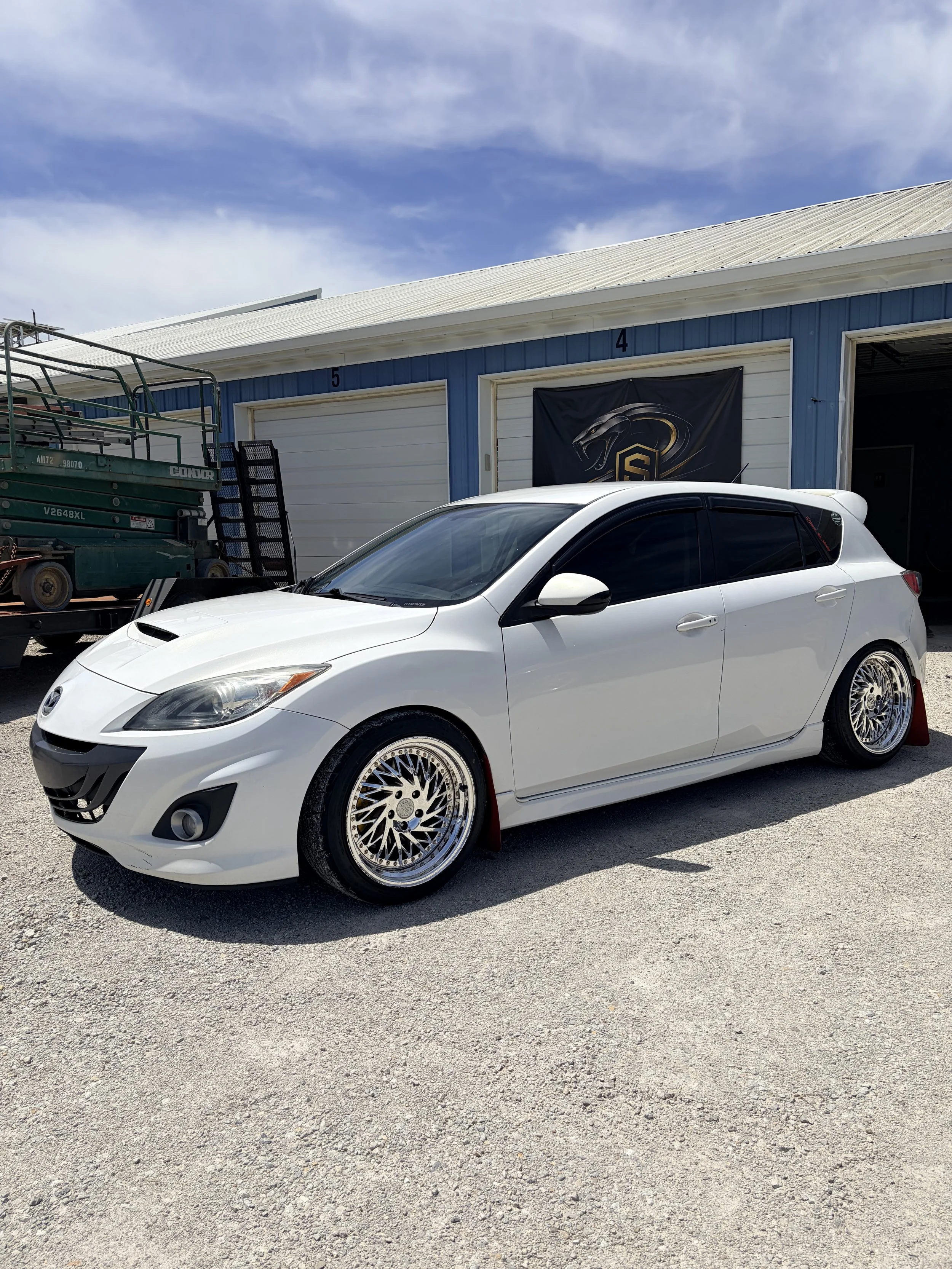 A white sports car with custom wheels parked in front of a blue garage with a logo of a panther on a black banner.