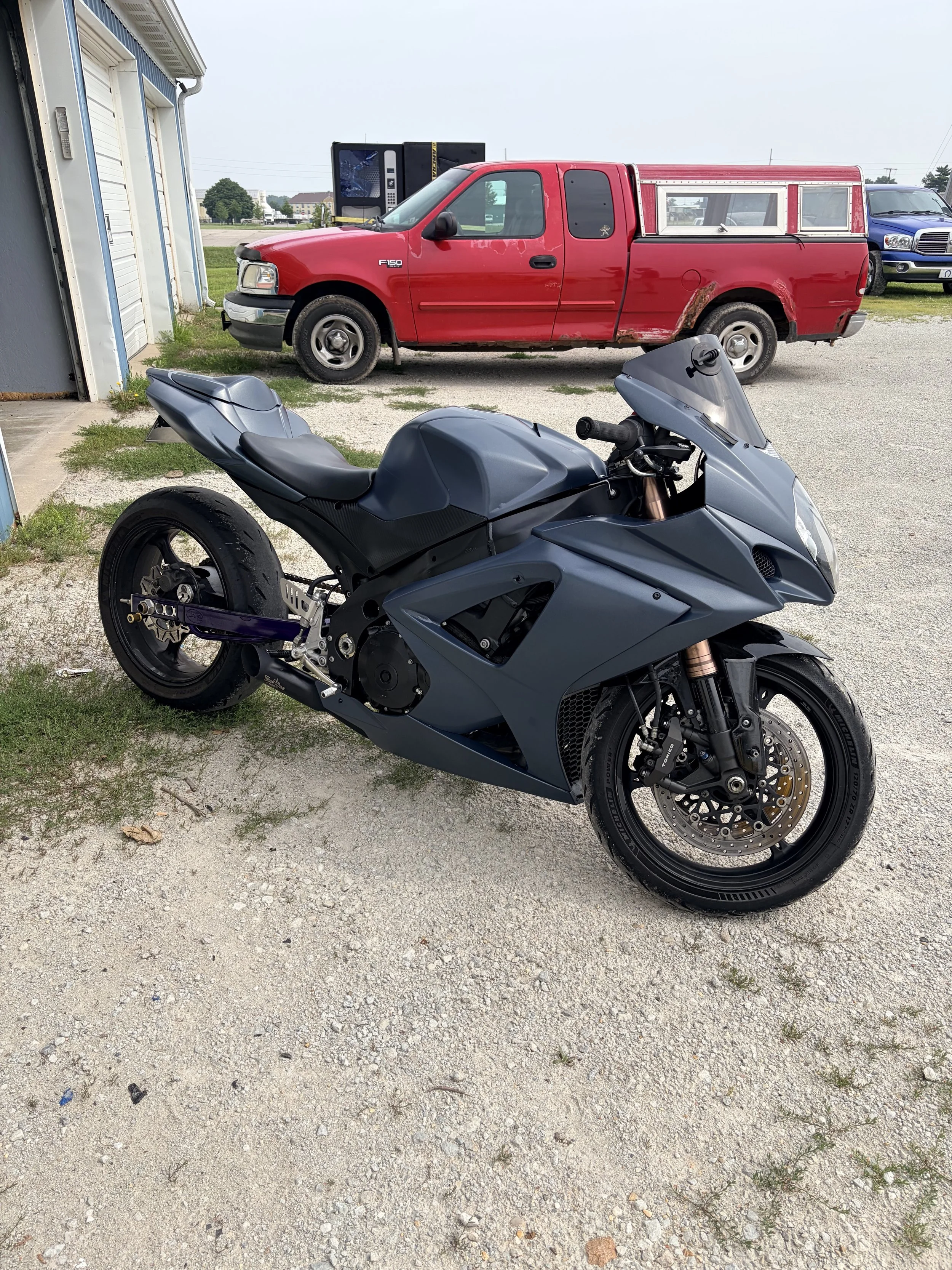 A black sport motorcycle is parked on a gravel lot with a red pickup truck and a blue vehicle in the background.