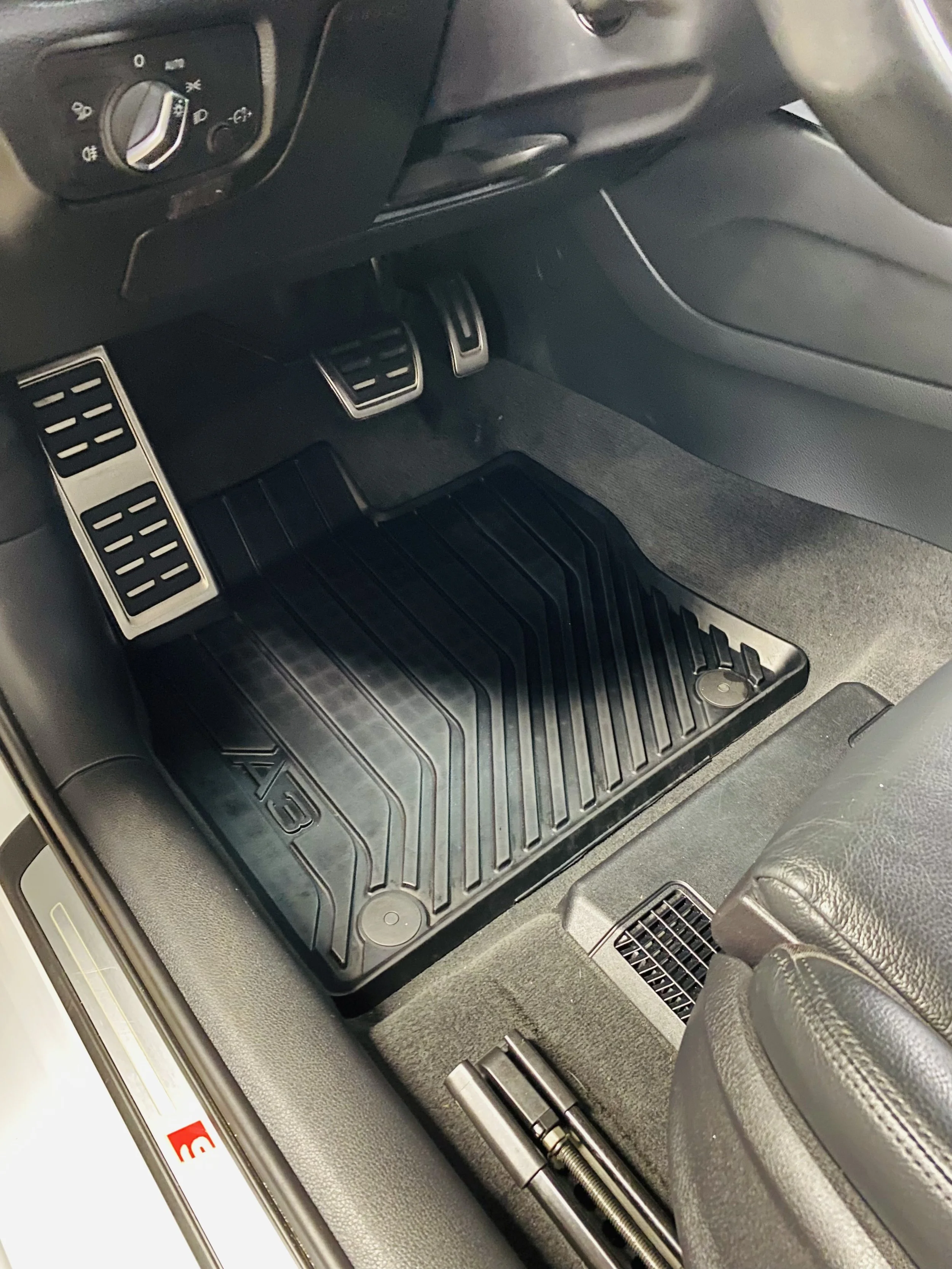 The image shows the interior of a car, focusing on the driver's side footwell. It includes accelerator, brake, and clutch pedals with metallic finishes, a black rubber floor mat, and a part of the car's dashboard with control knobs visible.