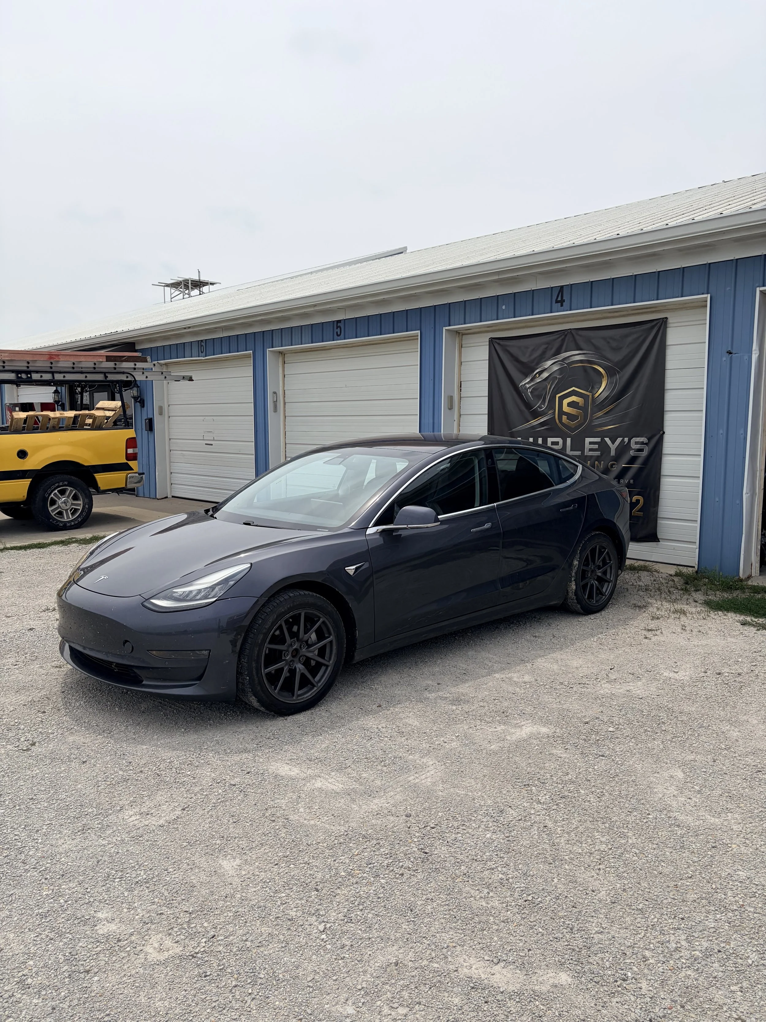 A black Tesla car parked in front of a Section 4 garage with a black and gold logo banner on the wall. There's a yellow truck with a ladder nearby, and a gravel surface under the vehicles.