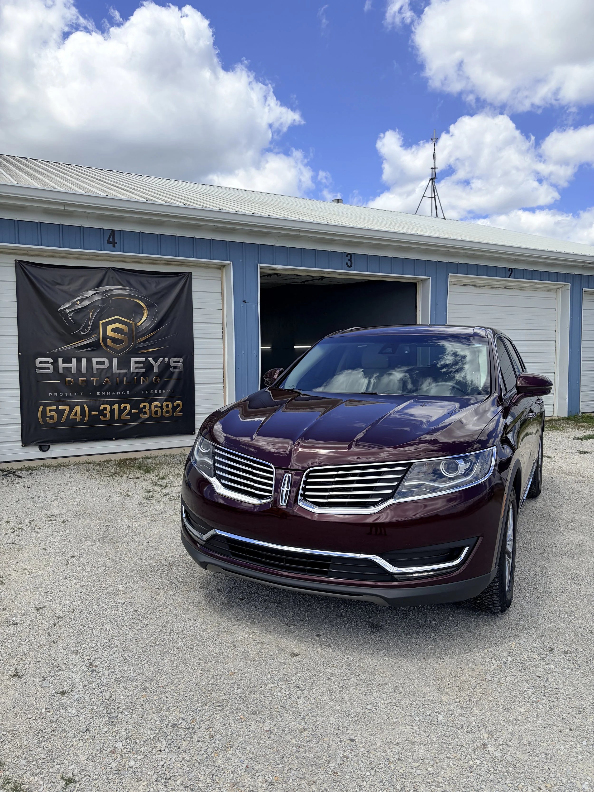 A shiny maroon car parked outside a blue and white building with a sign reading Shipley's Detailing. The building has numbered garage doors, and the sky is partly cloudy with a visible antenna on the roof.