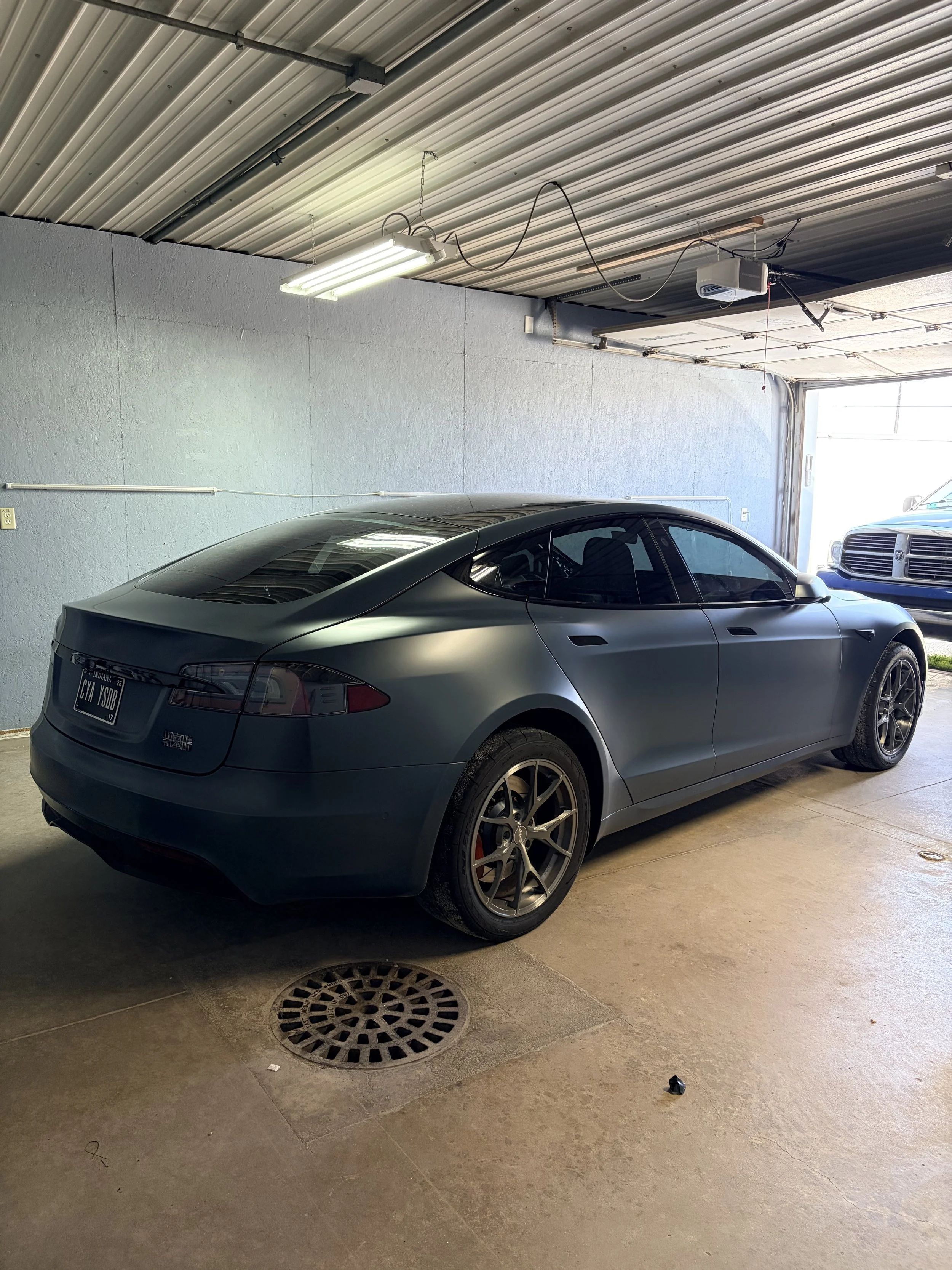 A gray sports car parked inside a garage with a concrete floor, gray wall, and a metal ceiling with hanging fluorescent lights and garage door opening. The cars have tinted windows and dark alloy wheels.