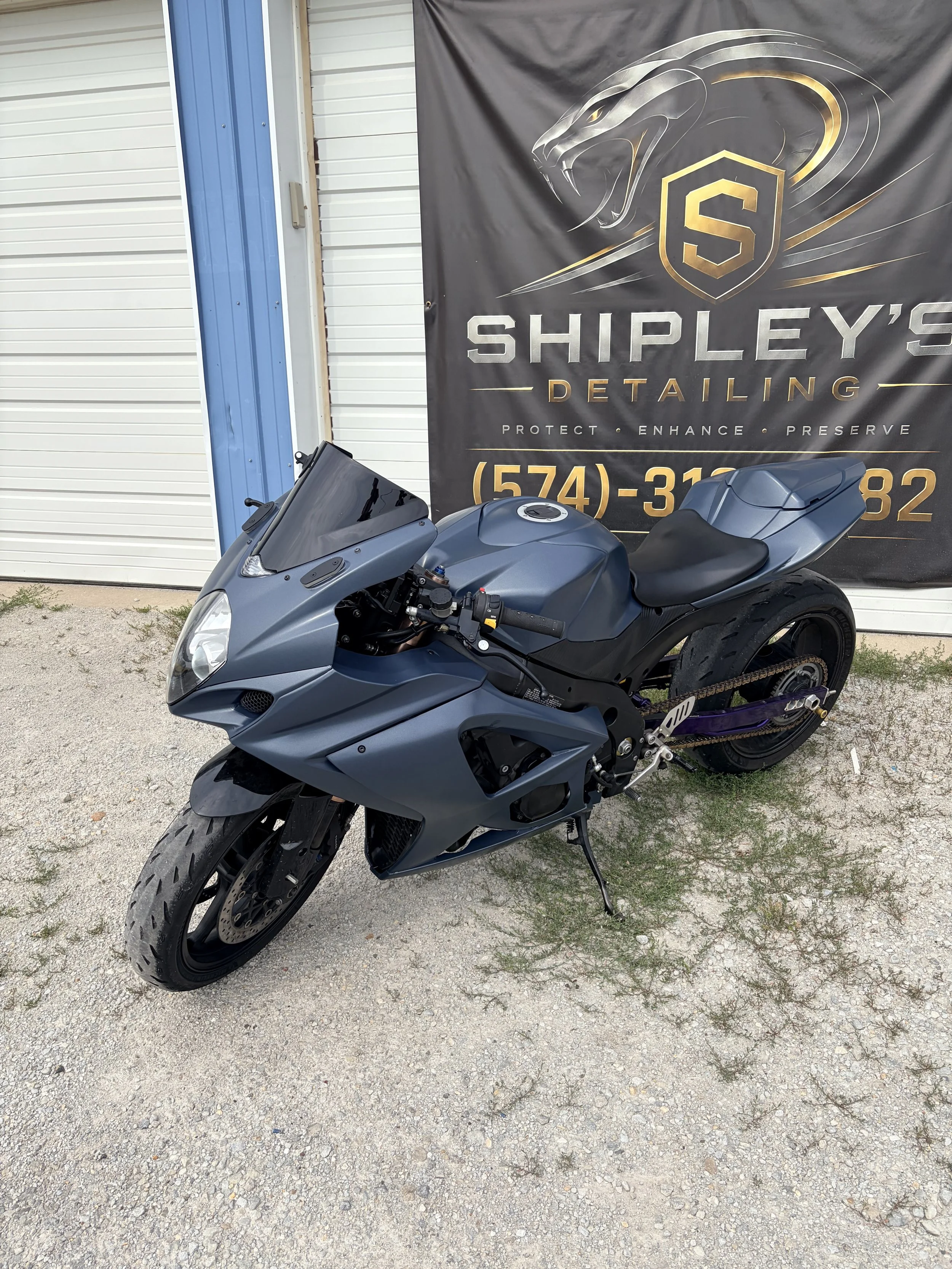 A gray sport motorcycle parked outdoors in front of a black banner with a lion logo and text for Shipley's Detailing, with a contact phone number.