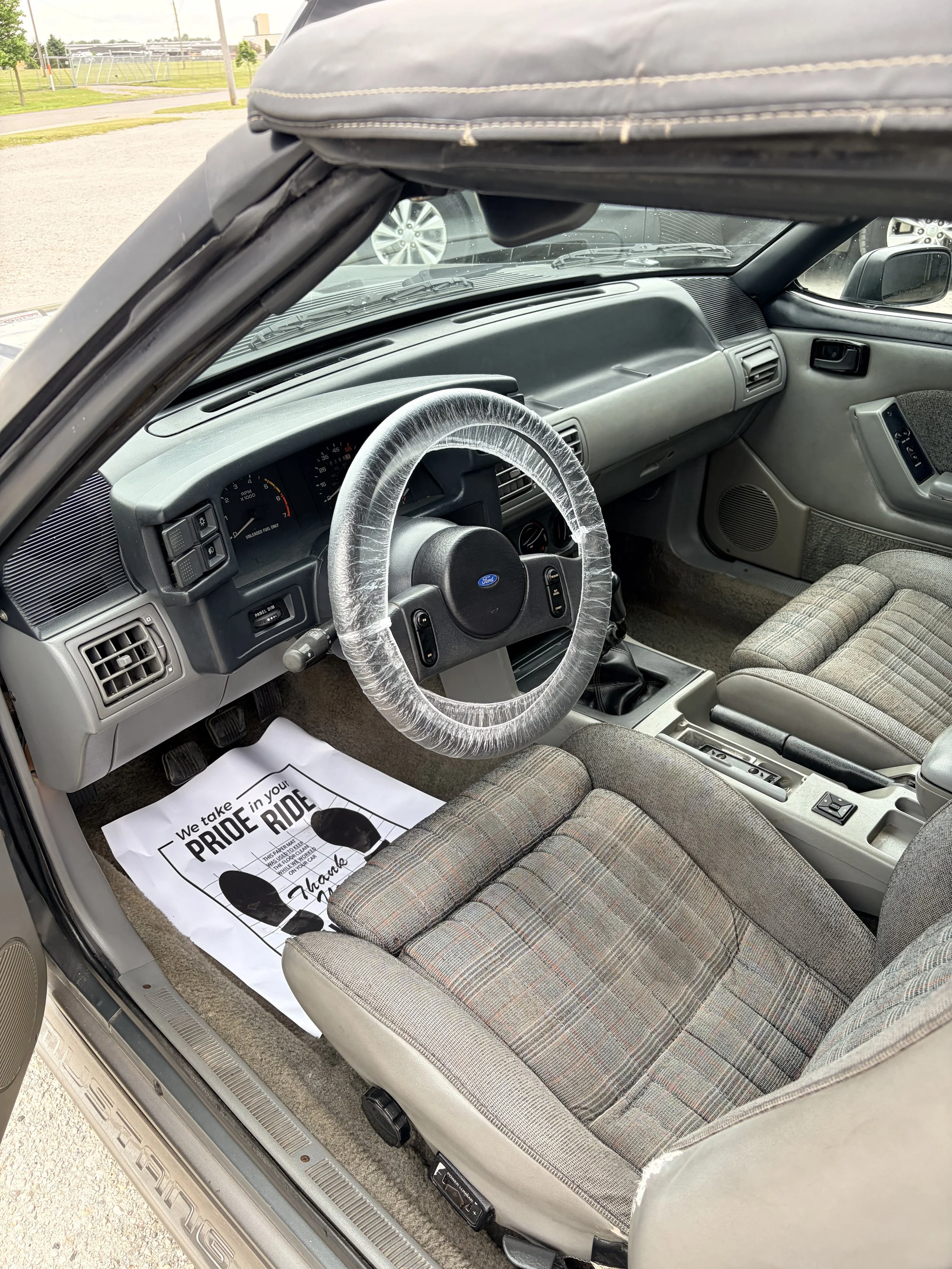 Inside an older model car, showing the front and passenger seats, steering wheel with a plastic cover, dashboard, and door panel. There is a promotional paper on the floor with a message about pride in riding.