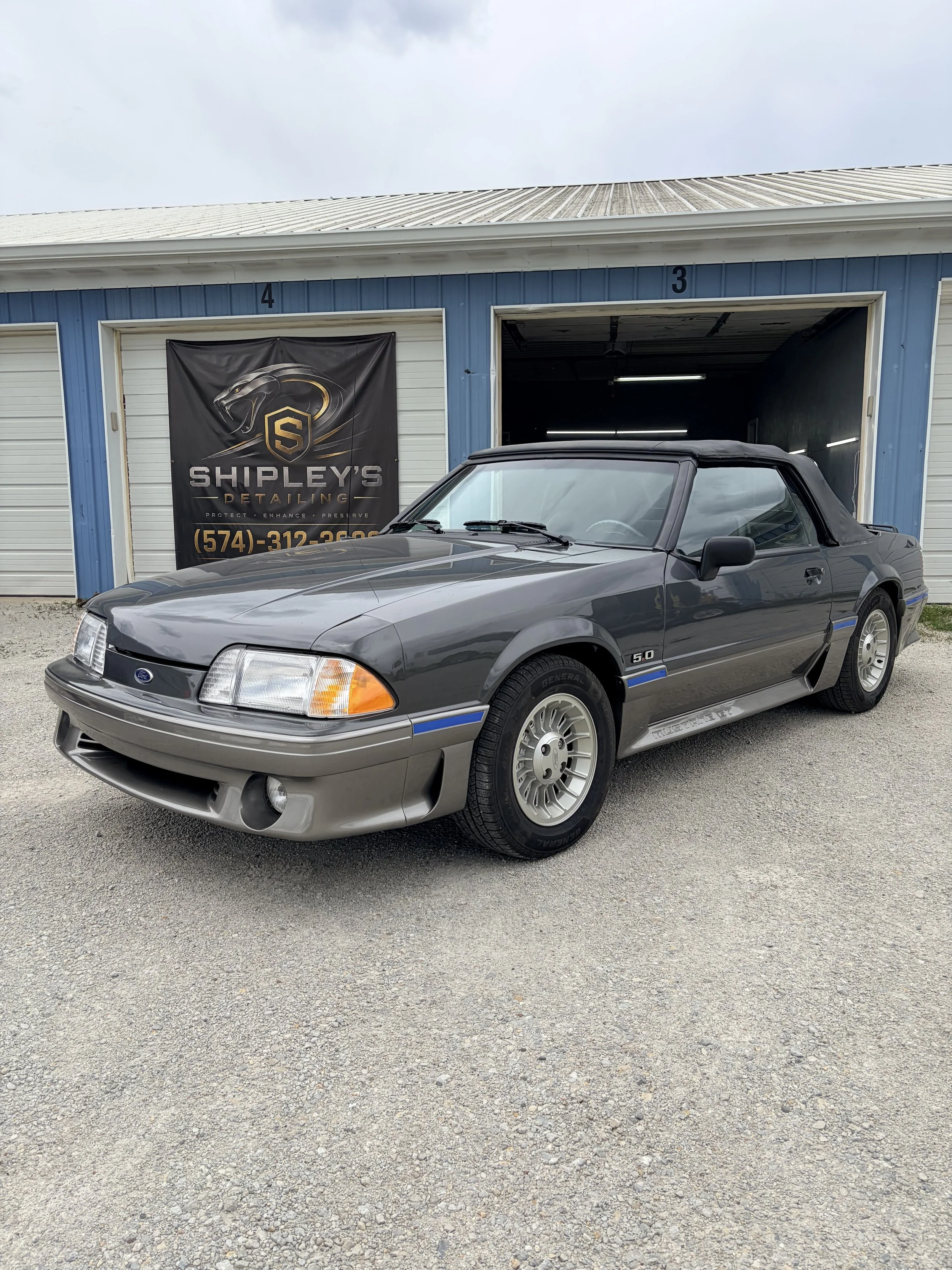 A gray Ford Mustang convertible with a black soft top parked outside a detailing shop with a blue building and a sign reading 'Shipley's Detailing'.