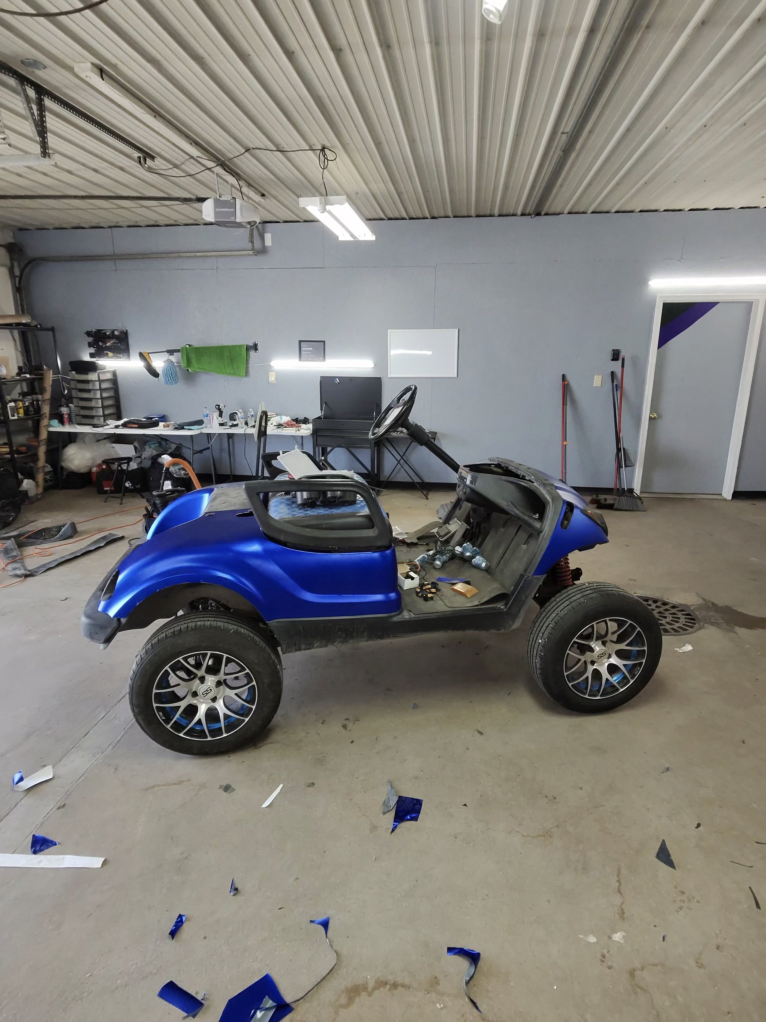 A partly assembled blue car inside a garage with a cluttered workbench, tools, and cleaning supplies. Confetti and debris on the concrete floor.