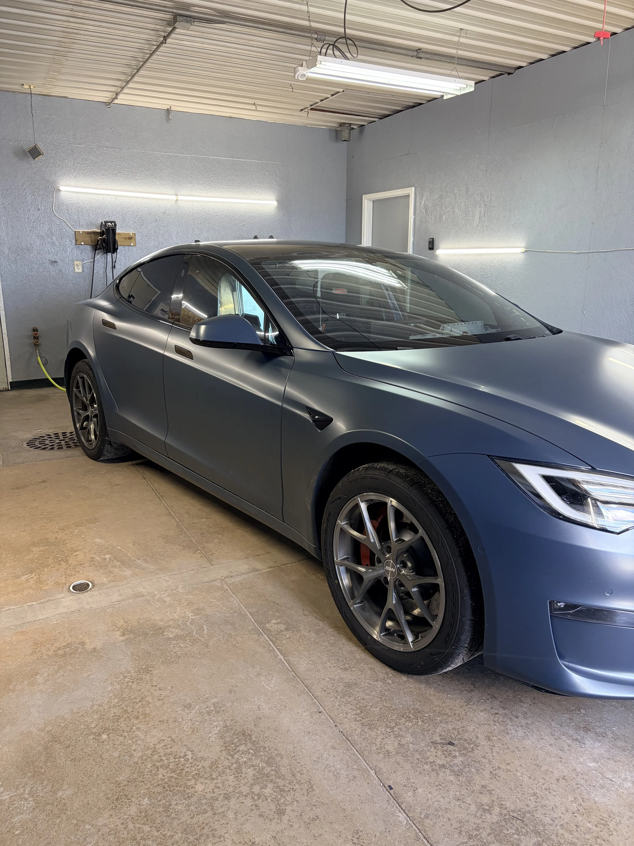 A sleek, metallic gray Tesla Model S parked in a garage with gray walls and a concrete floor. There are fluorescent lights on the ceiling and a wall-mounted power outlet with a charging cable connected to the car.