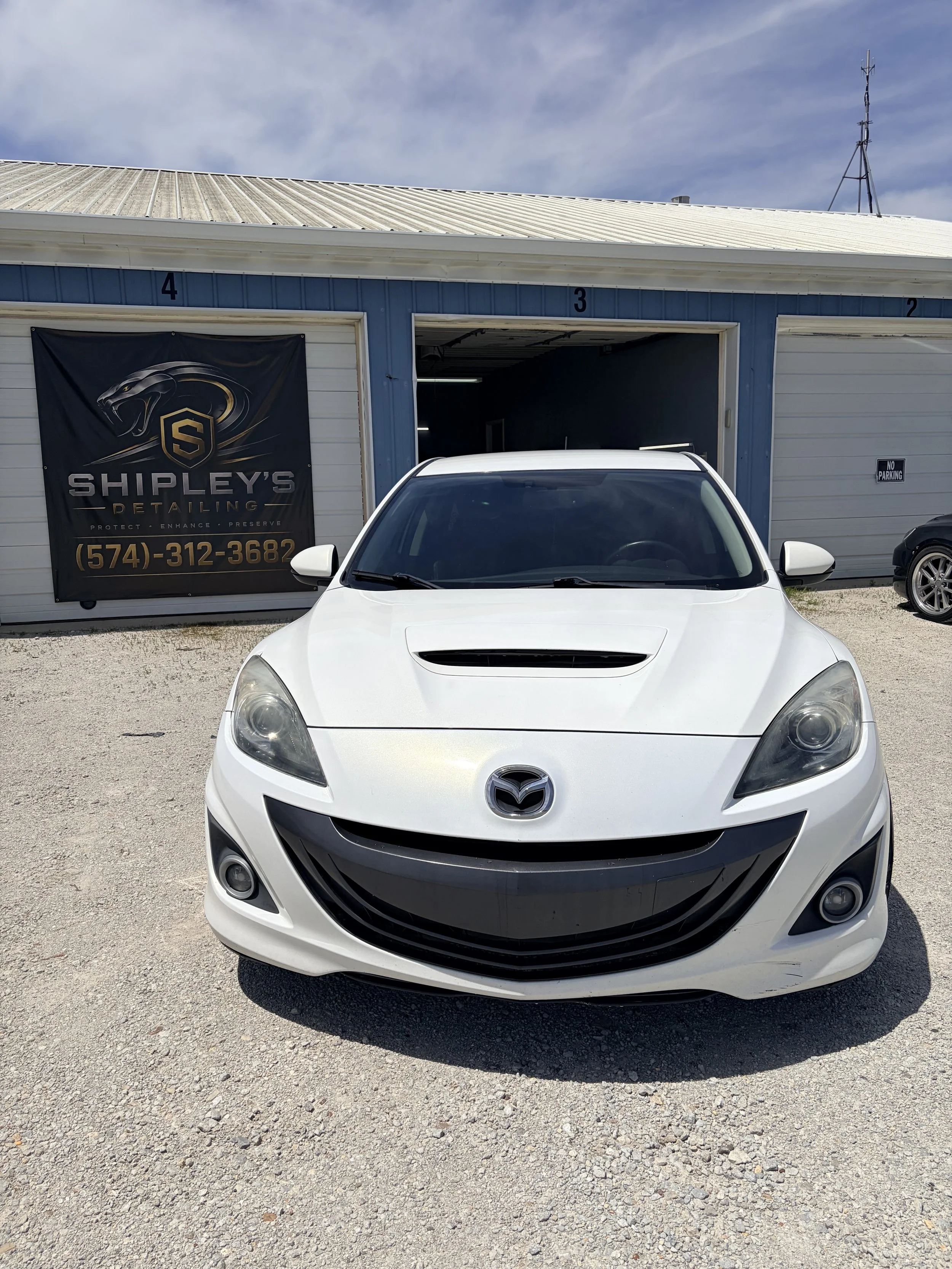 Front view of a white Mazda car parked outside an auto detailing shop with a black and gold banner, blue siding, large open garage doors numbered 4, 3, and 2, and a gravel ground.
