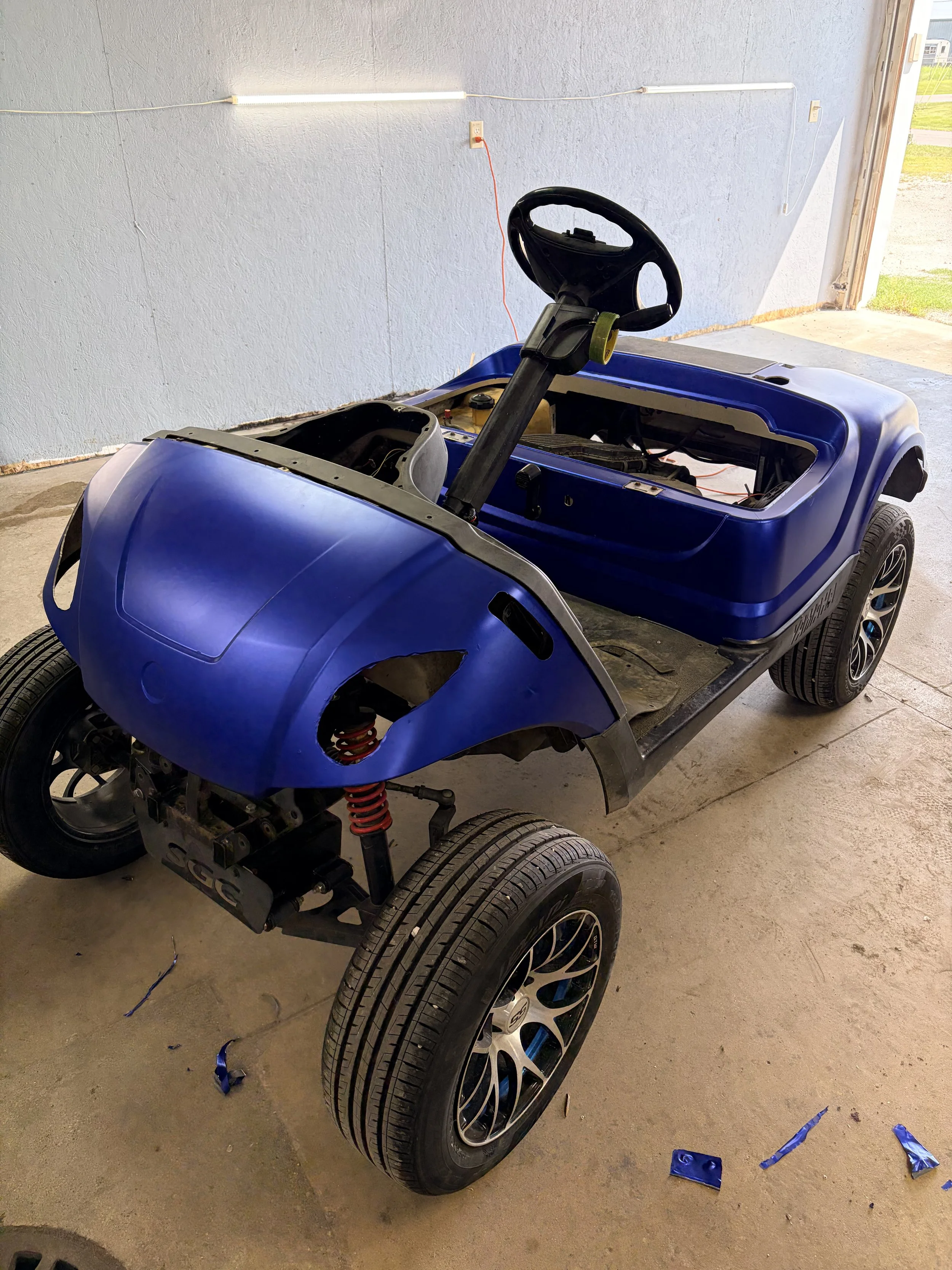 A blue golf cart with no seats, exposing dashboard and wiring, parked inside a garage with a concrete floor and a blue wall in the background.