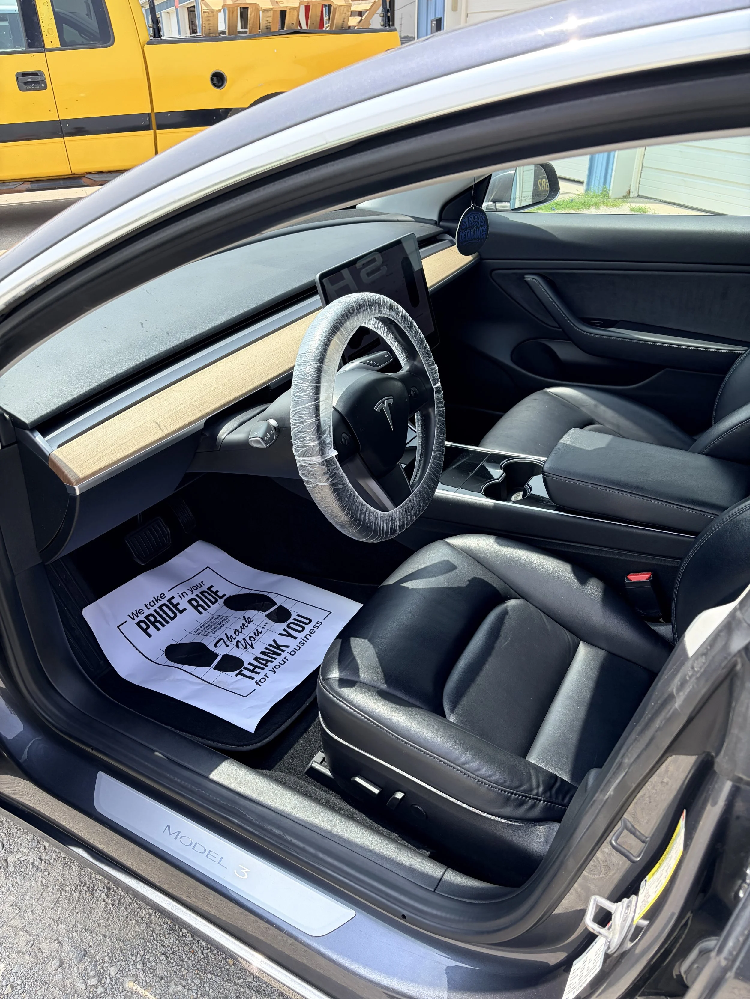 The interior of a Tesla Model 3 electric vehicle, featuring black leather seats, a wood accent dashboard, and a large digital display screen. A paper floor mat with a 