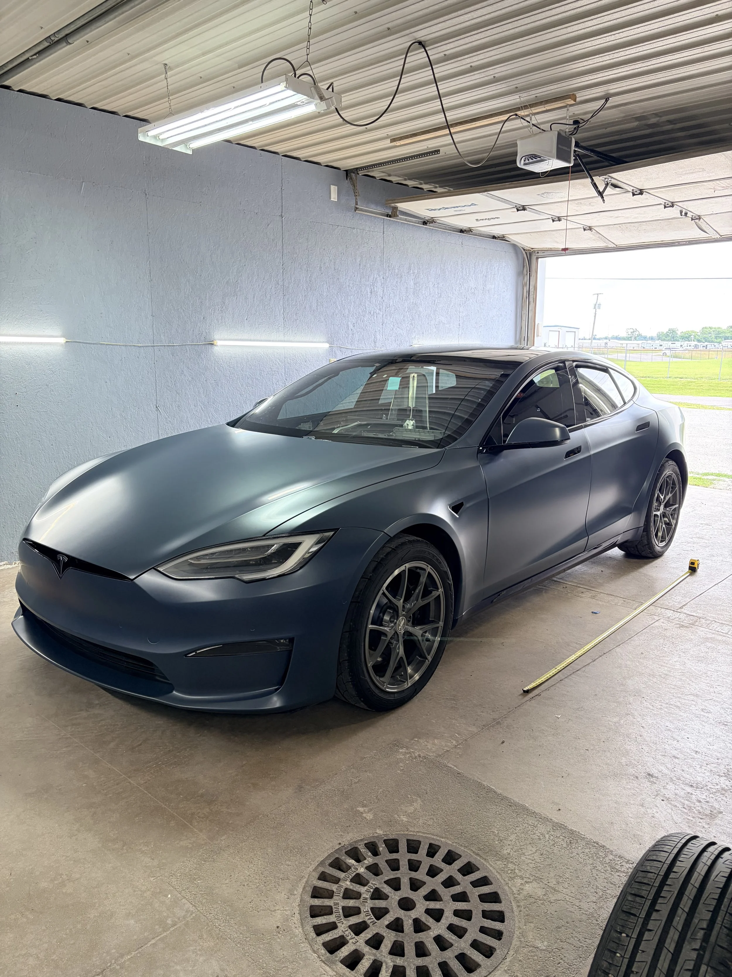 A gray Tesla sports car parked inside a garage with the garage door open, revealing a sunny outdoor area with grass and a fence.