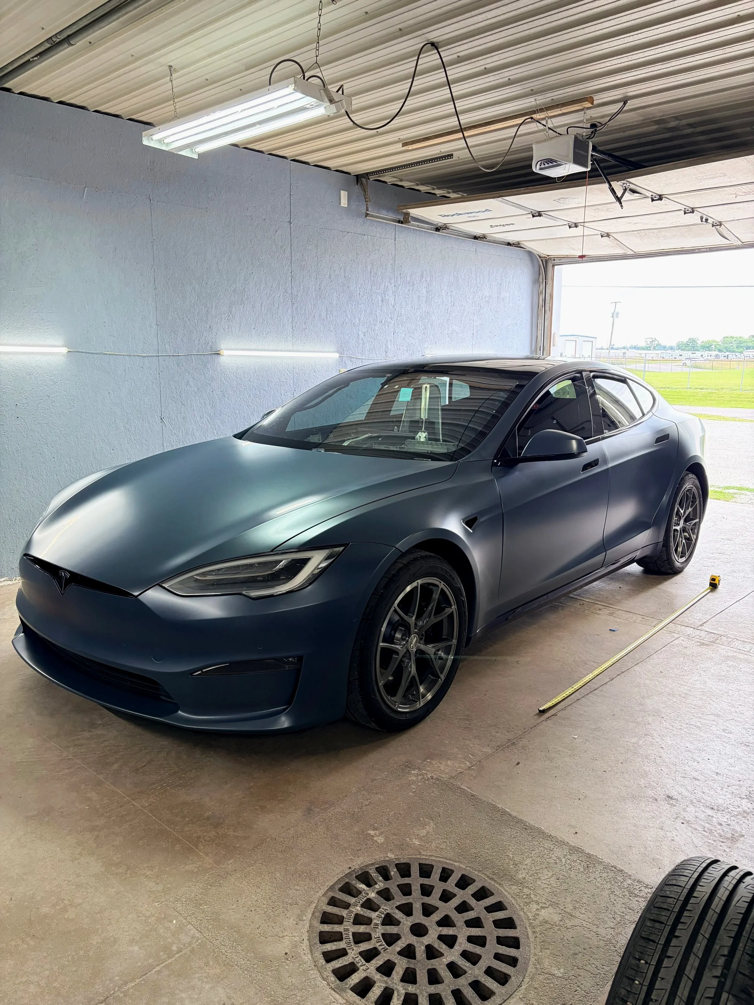 A blue Tesla sports car parked inside a garage with an open door. The garage has a corrugated metal ceiling with fluorescent lights and a wall with a light blue painted surface. There is a measuring tape on the floor, and two tires placed in the bottom right corner of the image.